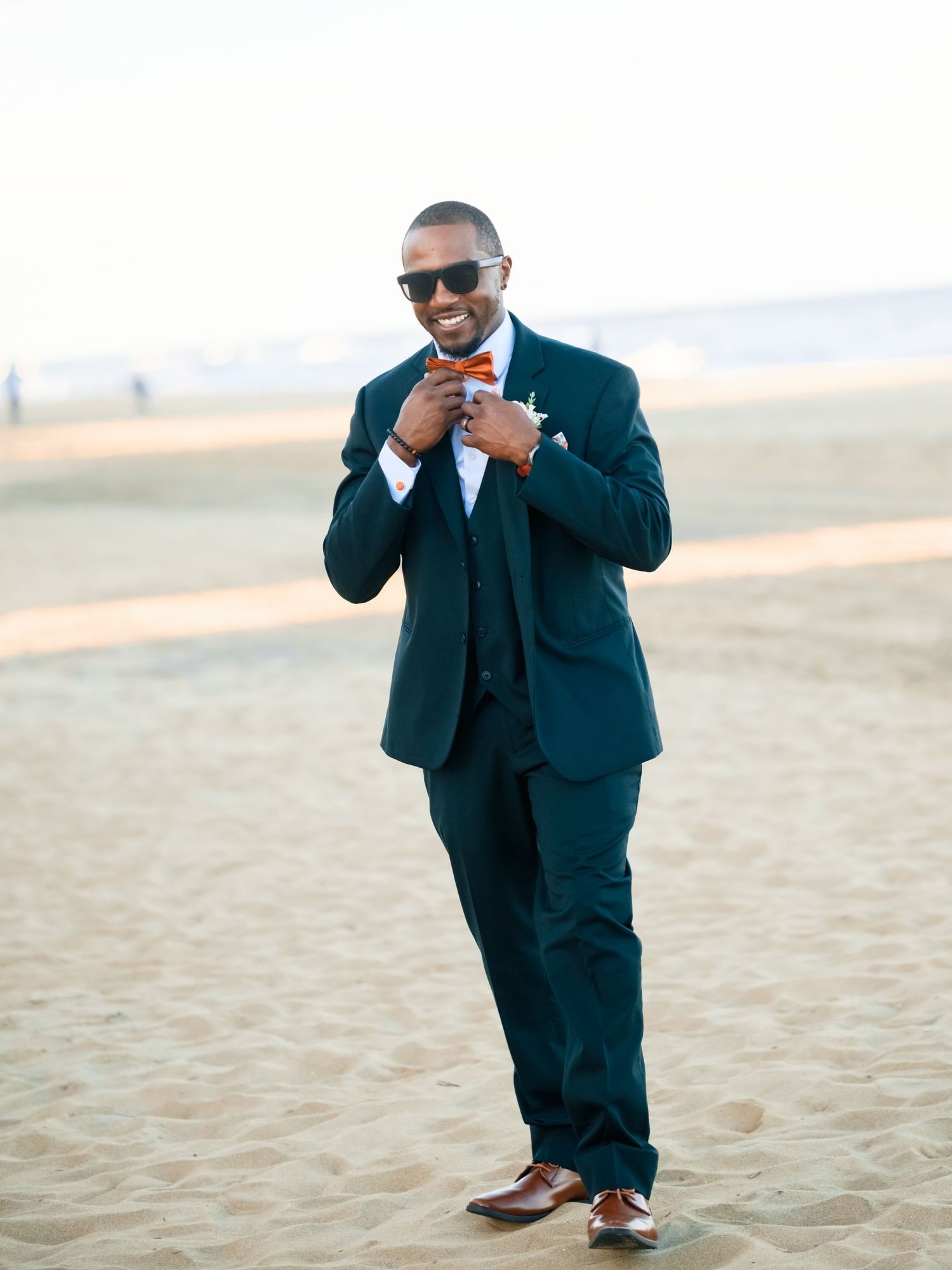 A person in a black tuxedo poses playfully on the beach sand.