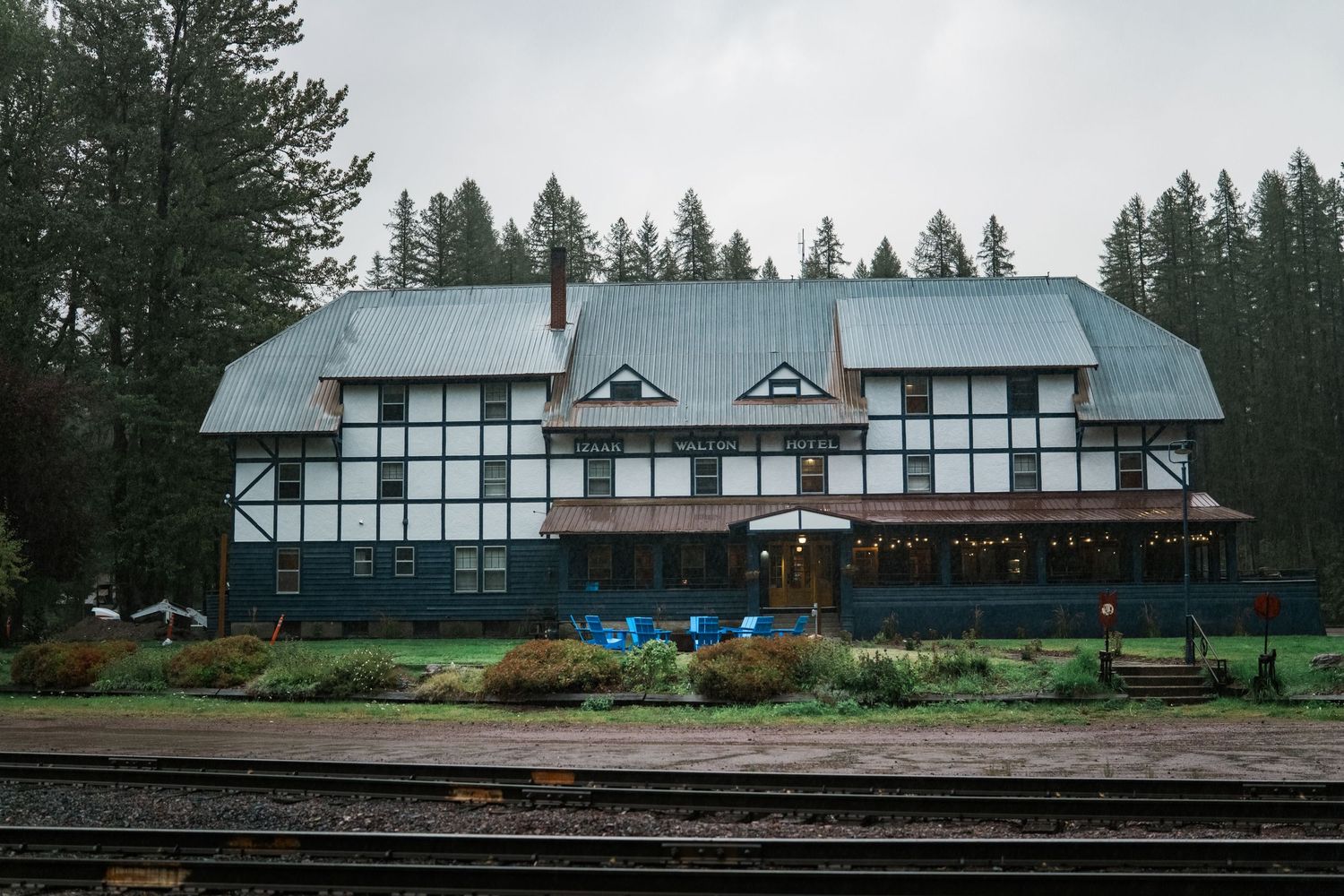 A panoramic view of a rustic lodge with checkered windows nestled among evergreen trees along railroad tracks.
