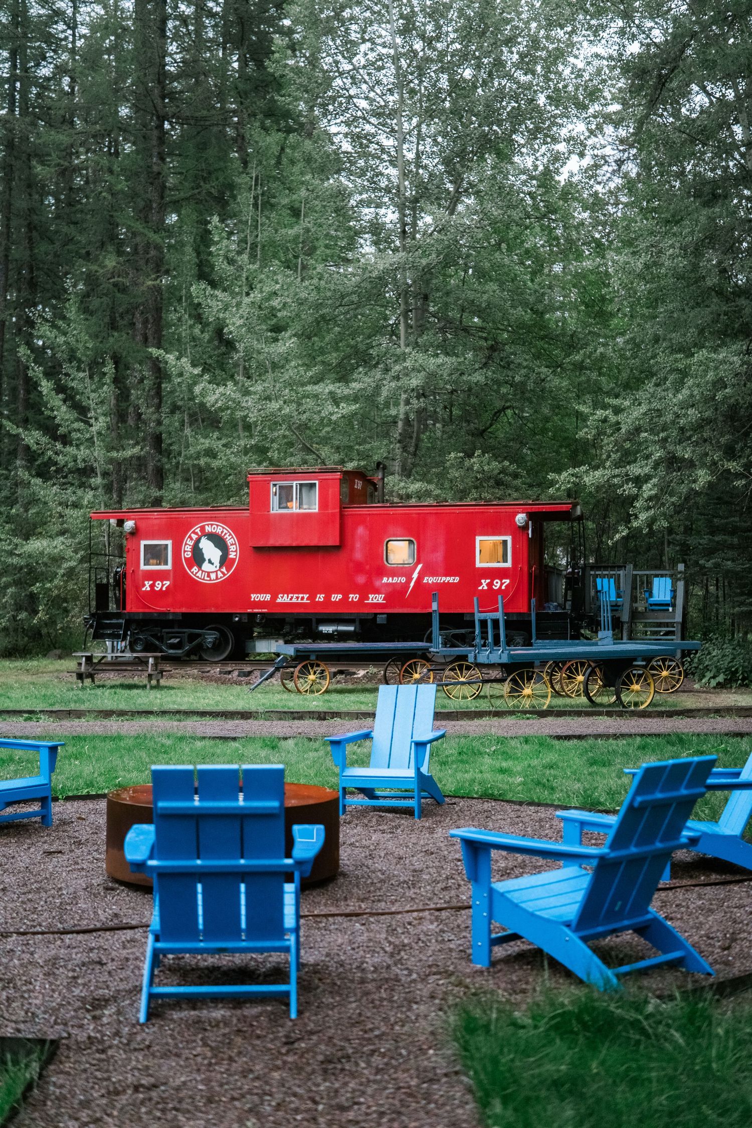 A red caboose train car displayed next to blue picnic benches in a wooded outdoor setting provides a scenic backdrop.