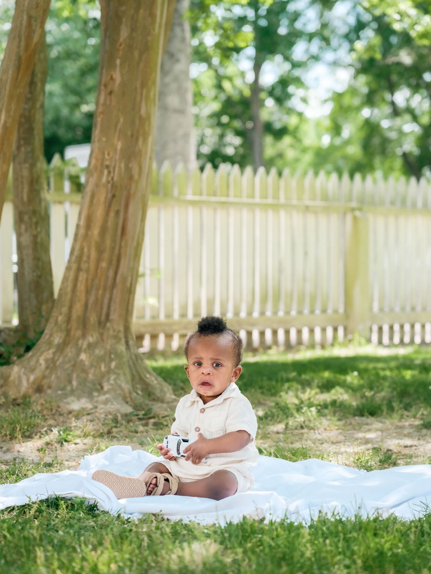 Baby lies on a white blanket outdoors with a wooden picket fence and trees in the background.