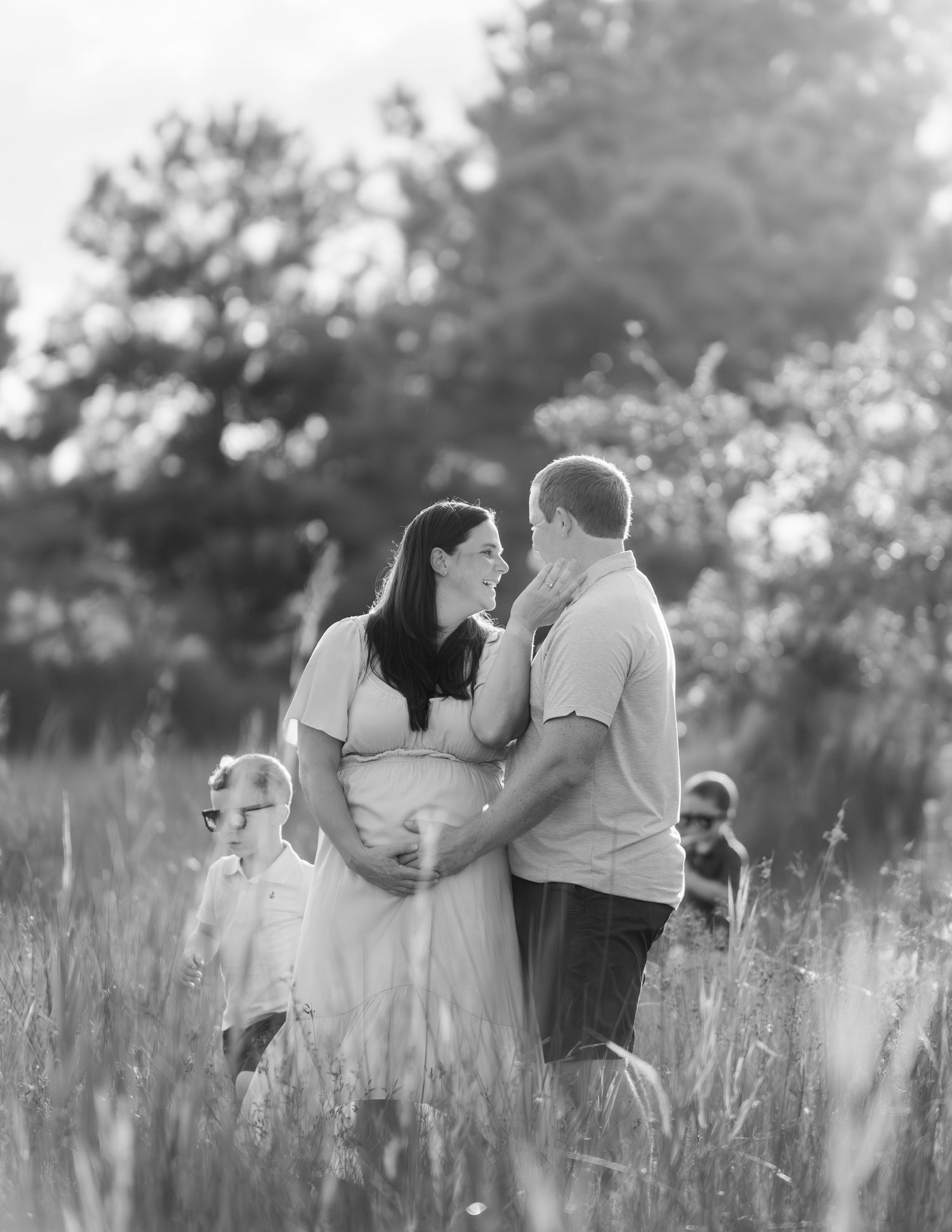 A black and white family maternity photo captured in tall grass at sunset with dreamy backlighting and soft bokeh.