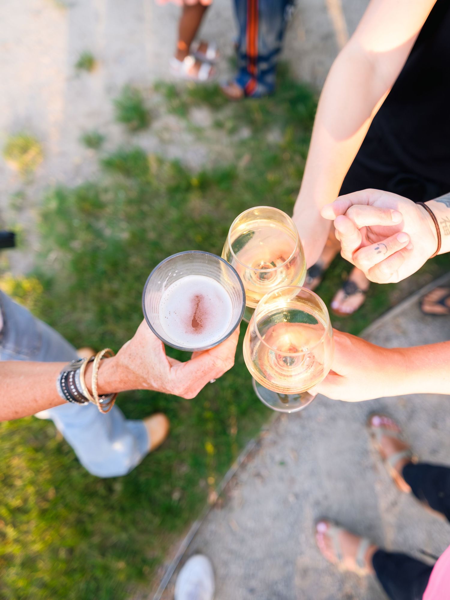 Friends toasting with various beverages outdoors on a sunny day, showing hands holding glasses in celebration.