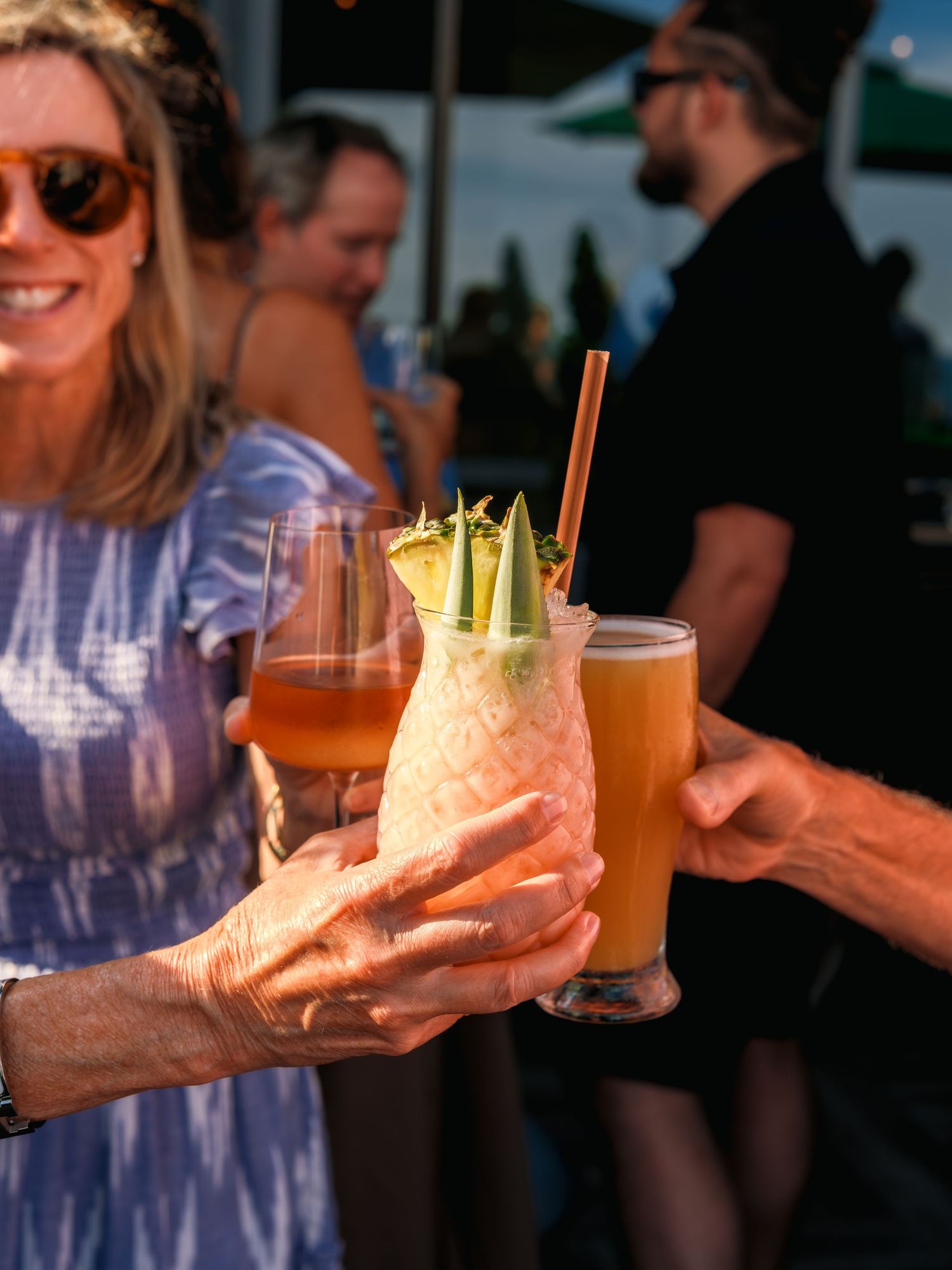 People holding and cheering with vibrant cocktail drinks at a summer outdoor party during golden hour.