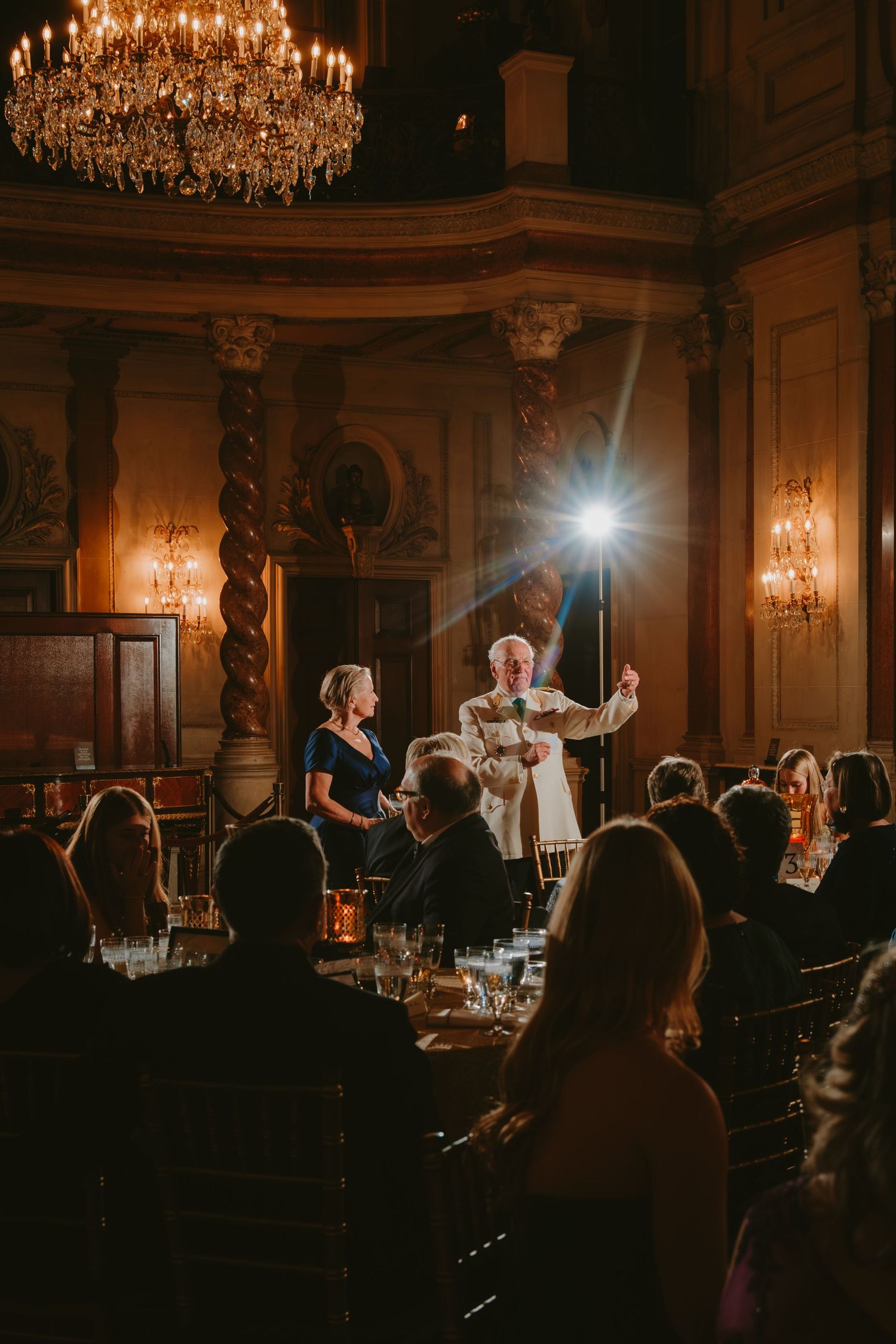 Elegant ballroom event with crystal chandelier and wall sconces illuminating guests gathered for an evening celebration.