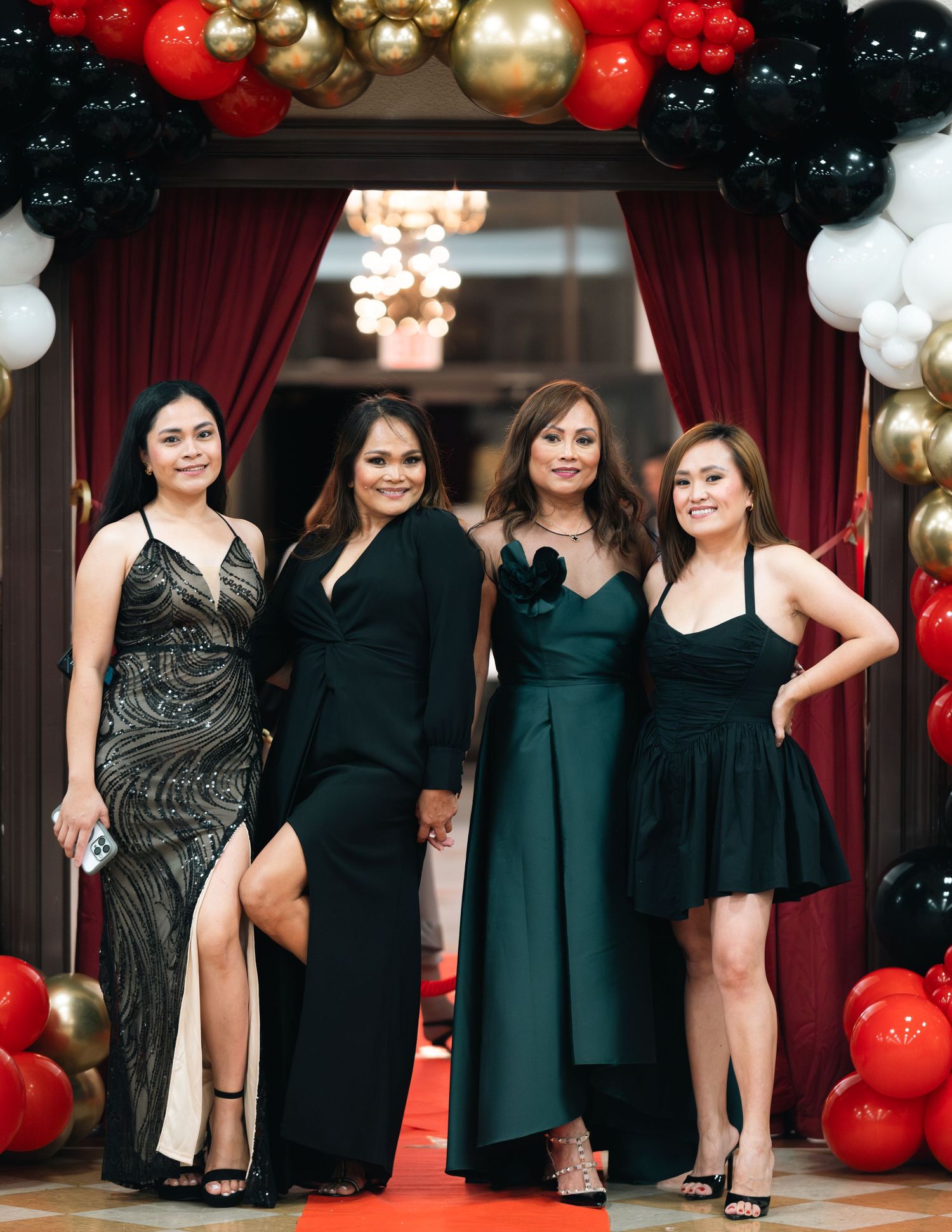 A formal event with red, gold, and black balloon arch decor and burgundy curtains framing guests in elegant dark dresses.