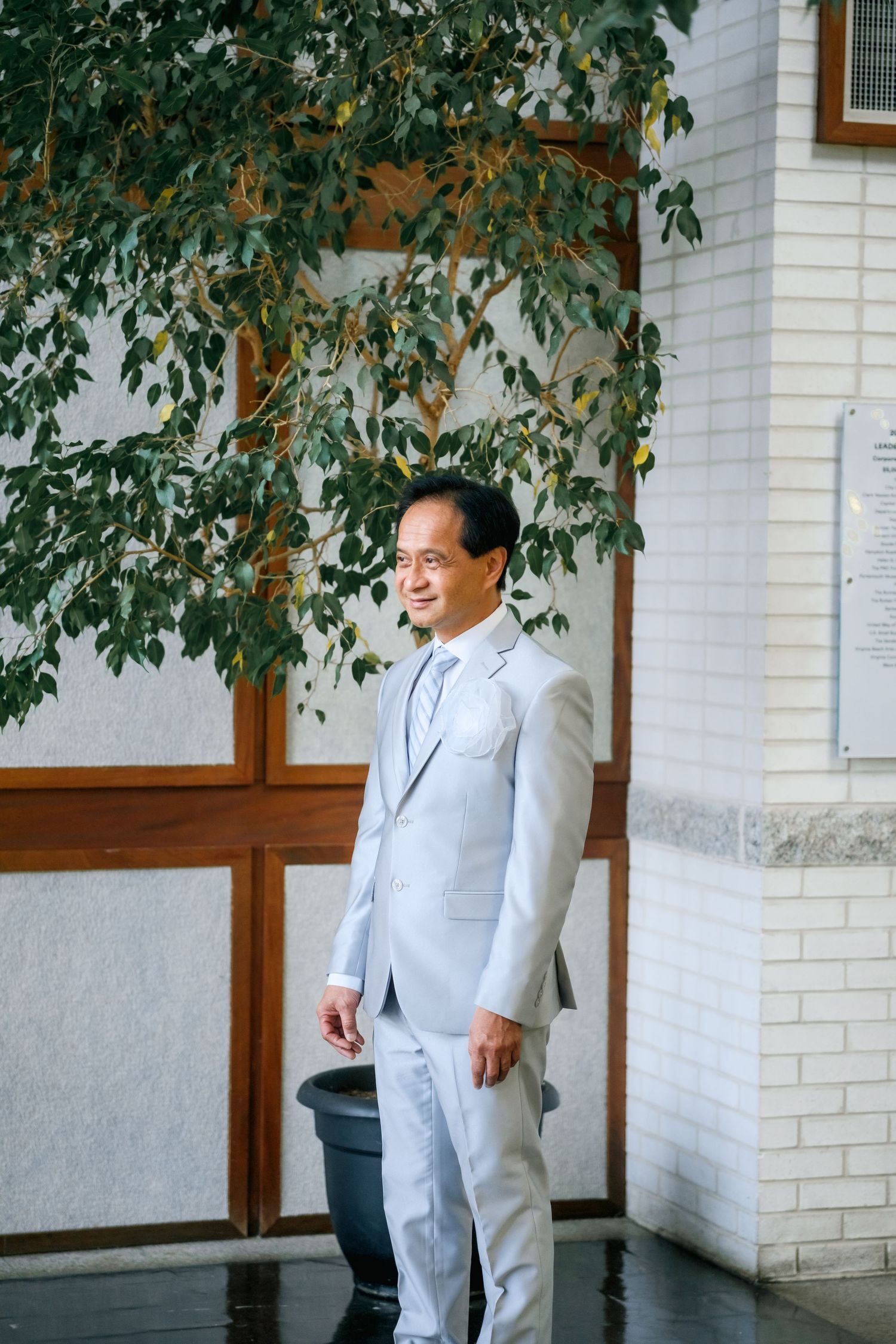 Professional in a light colored suit smiles while standing against a white brick wall with green foliage above.