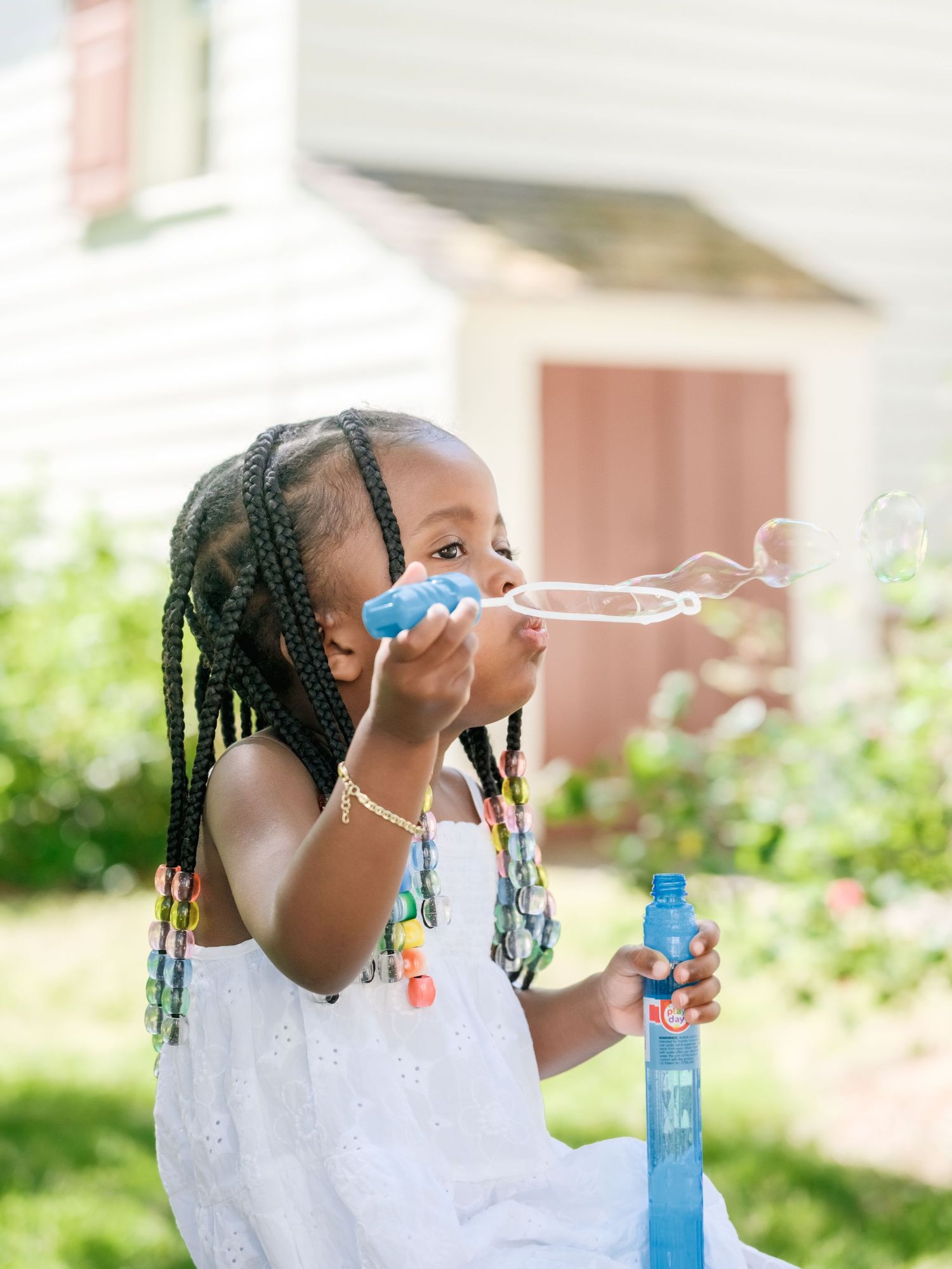 Young one in a white dress plays with soap bubbles in a sunny garden setting while holding a bubble wand.