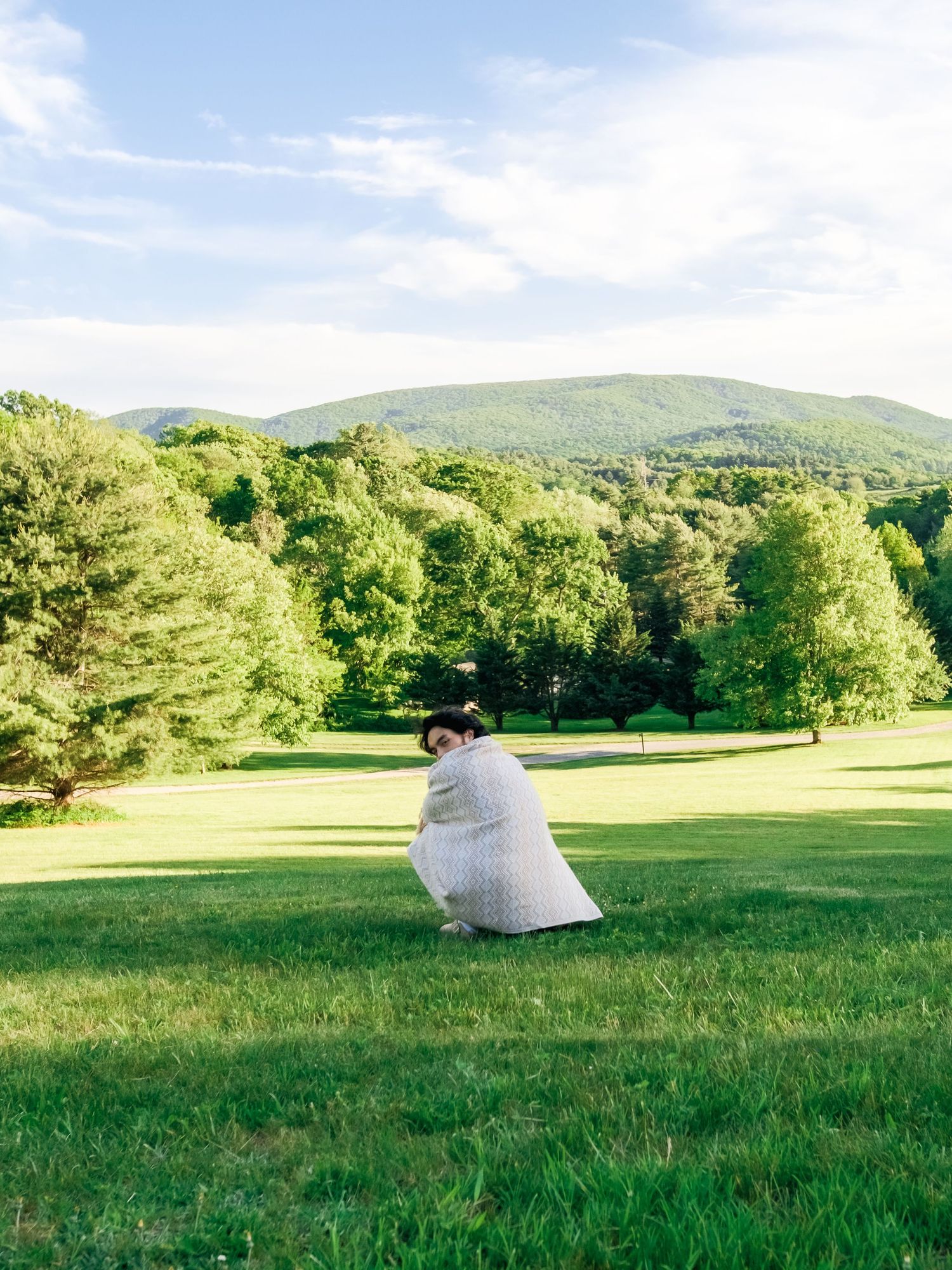 Person in white clothing sits in a peaceful green field with rolling hills and trees in the background on a sunny day.