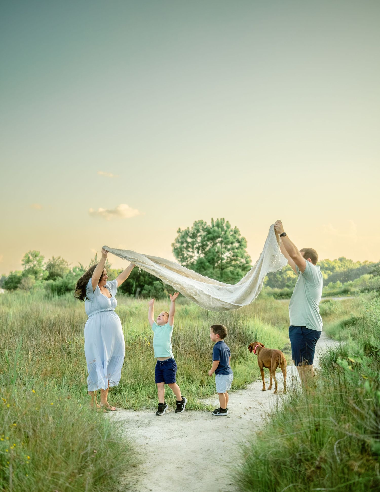 A family and their dog play with a flowing white sheet on a nature trail at sunset in a dreamy outdoor setting.