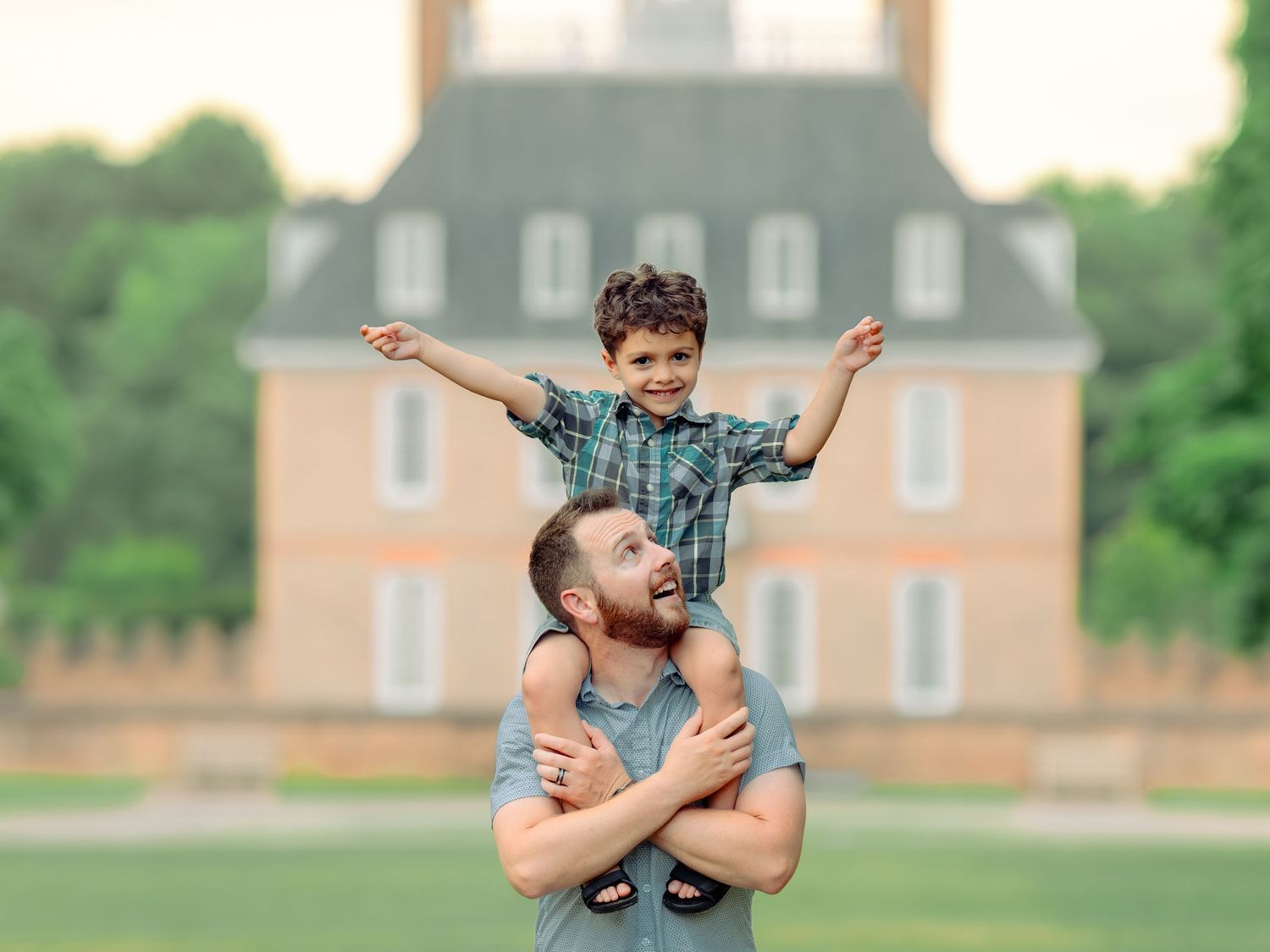 A father and son share a joyful moment playing outdoors in front of a large brick house during summer.