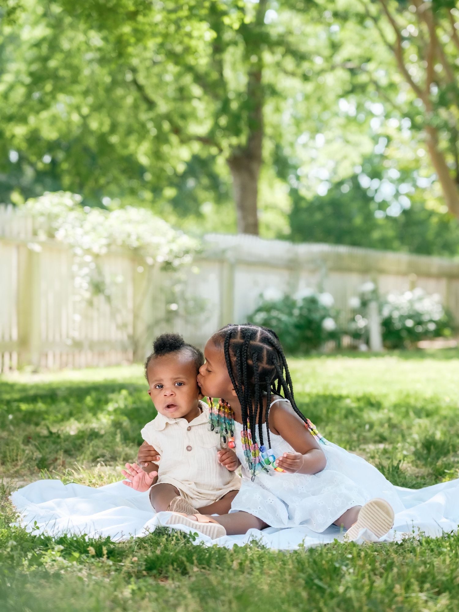 Two children sitting on a white blanket in a sunny backyard with a white fence and greenery in the background.