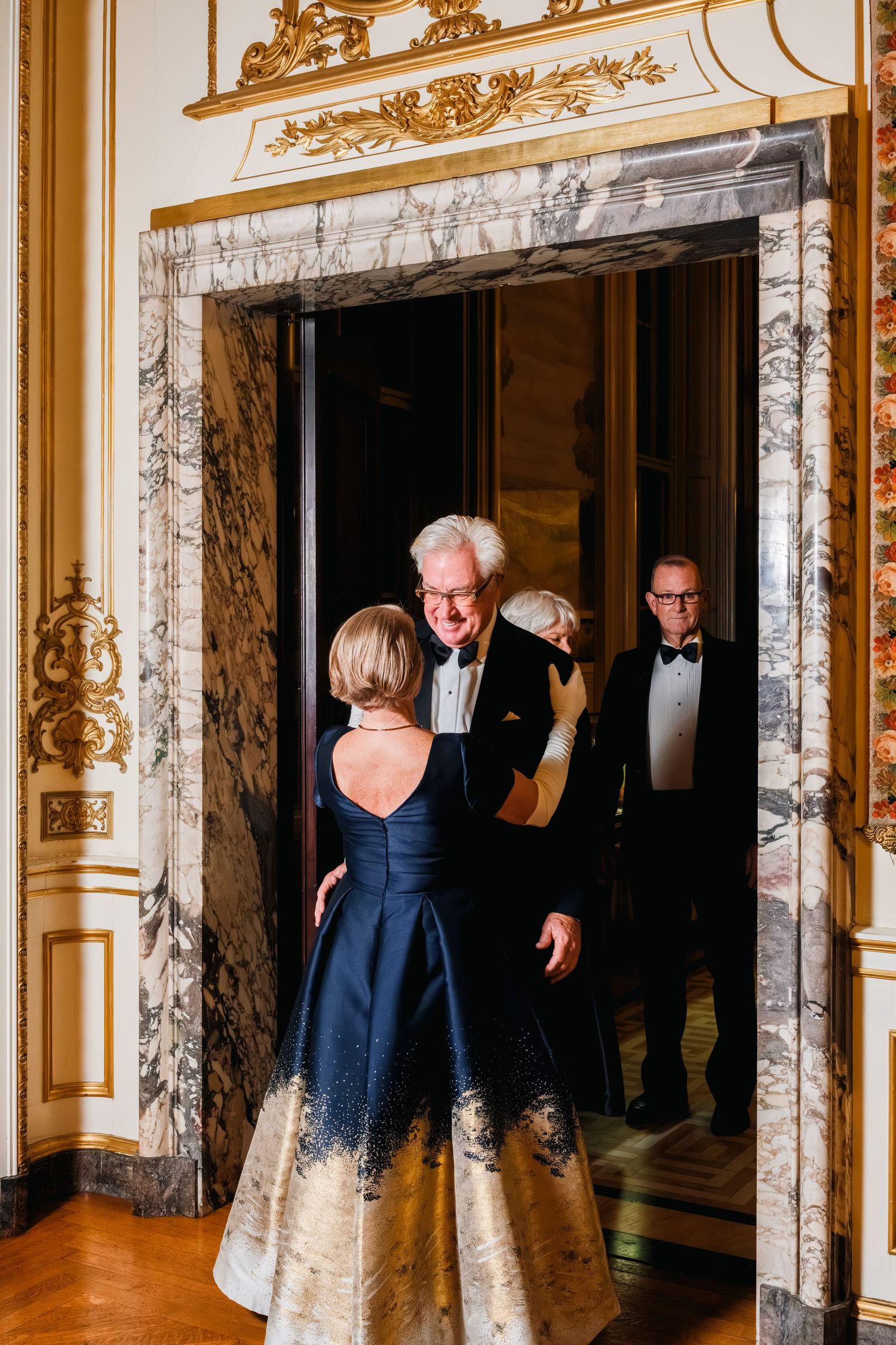 Elegant ballroom with ornate gold trim and marble doorways shows guests mingling at a formal event.