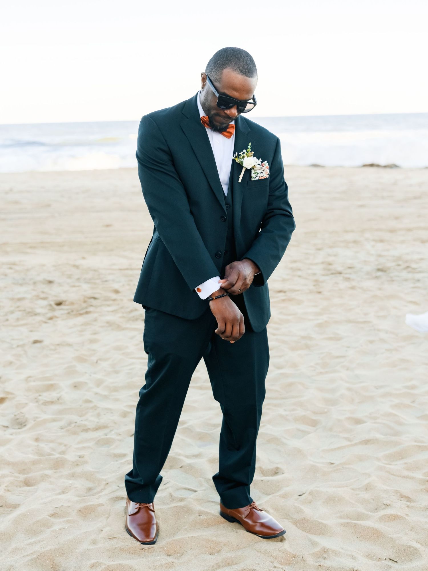 A formal wedding party member in a dark suit and bow tie poses on a sandy beach.