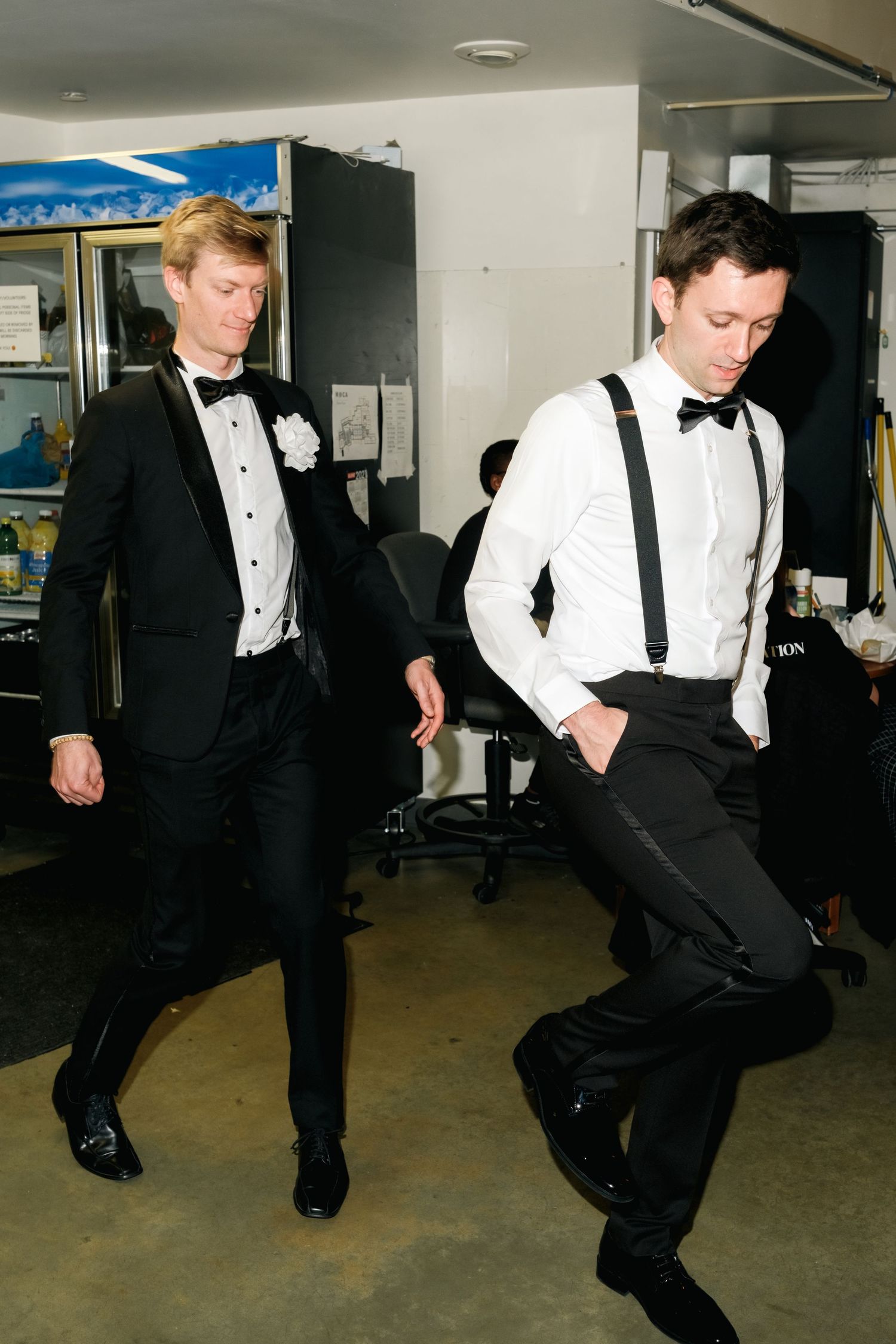 Two men in formal black tie attire with bow ties and suspenders dance in what appears to be a backstage area.