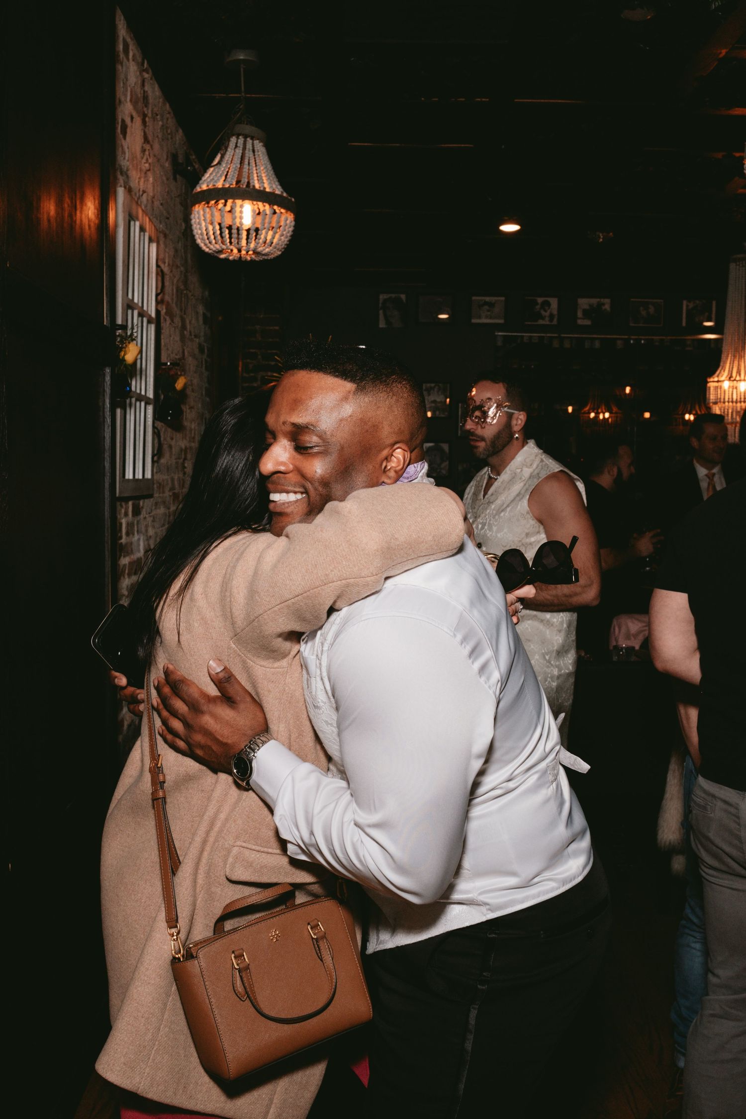 Two people share a warm embrace at a dimly lit indoor venue with crystal chandeliers overhead.