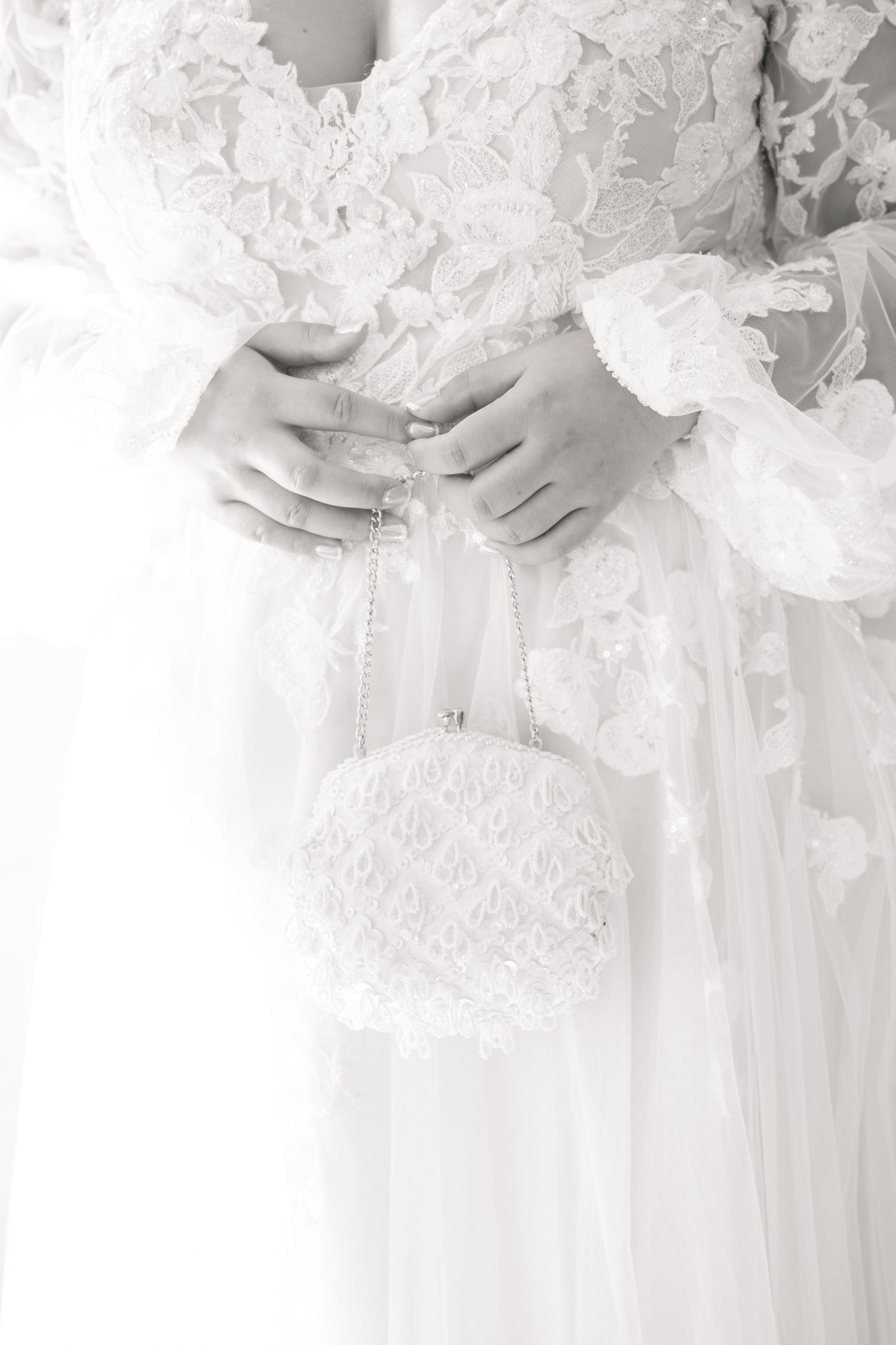 A delicate beaded white bridal purse hangs from hands wearing lace sleeves in a soft black and white photo.