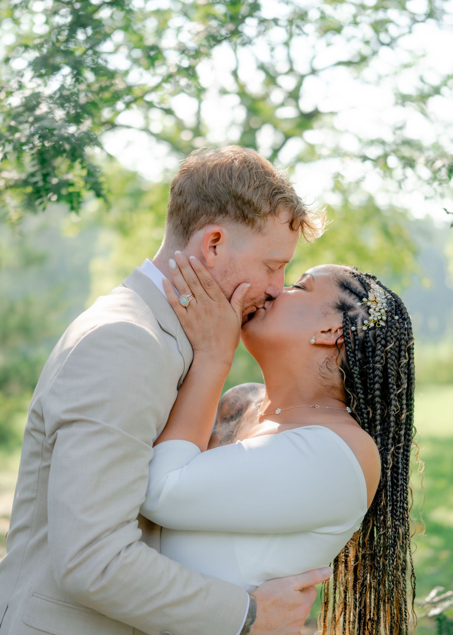 A couple shares a romantic kiss in a garden setting while wearing wedding attire against a soft natural backdrop.