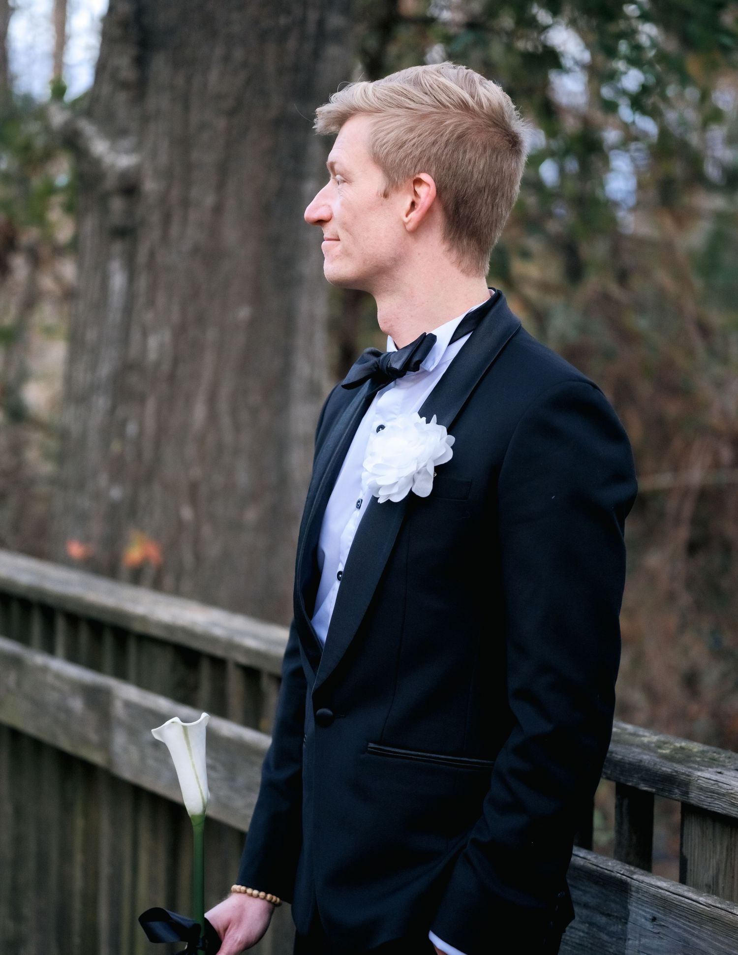 A groom in a black tuxedo and white boutonniere gazes contemplatively at the scenic view from a wooden deck.