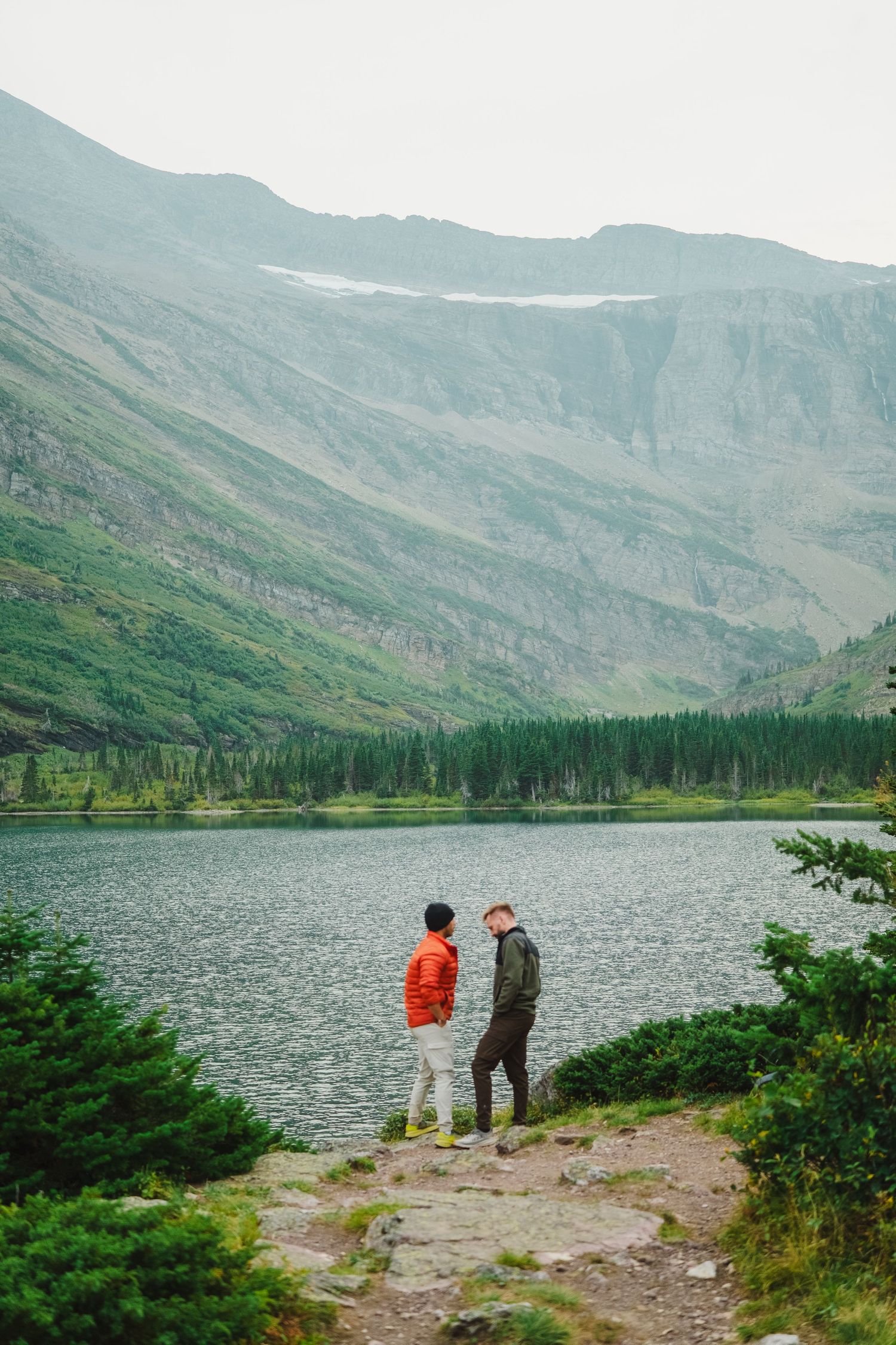 Two people in jackets stand at the rocky edge of a pristine mountain lake surrounded by evergreen forest and peaks.