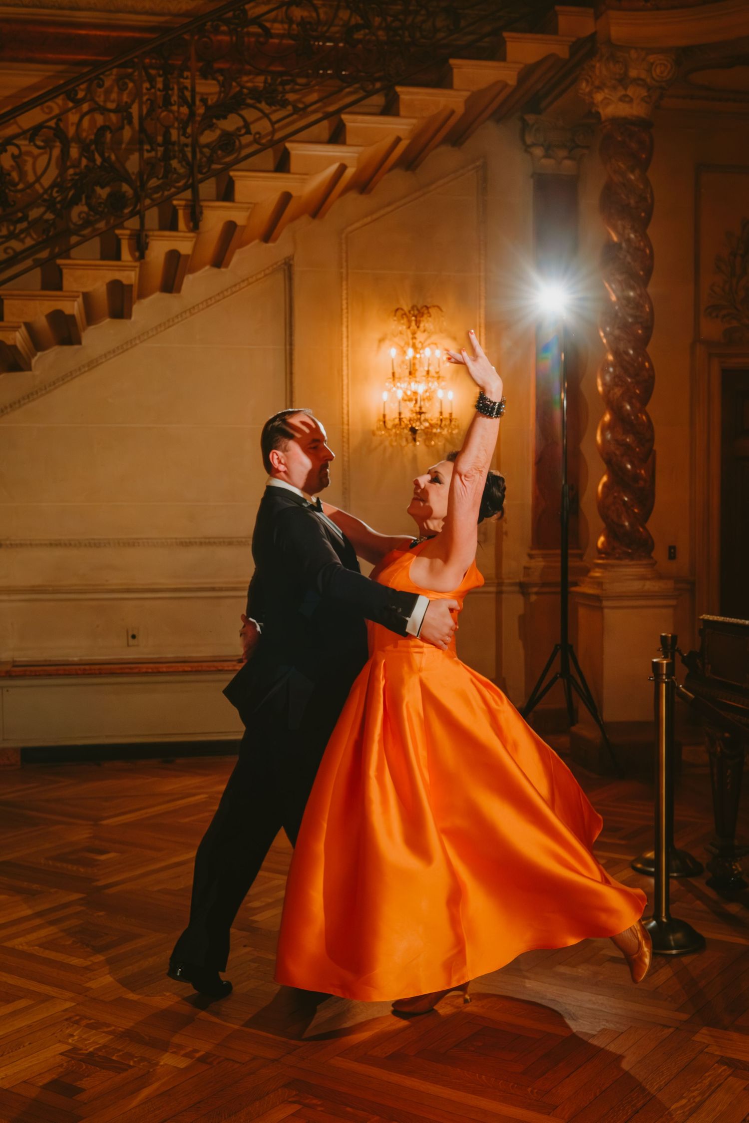A couple dances elegantly in a grand ballroom, with the woman wearing a vibrant orange ball gown.