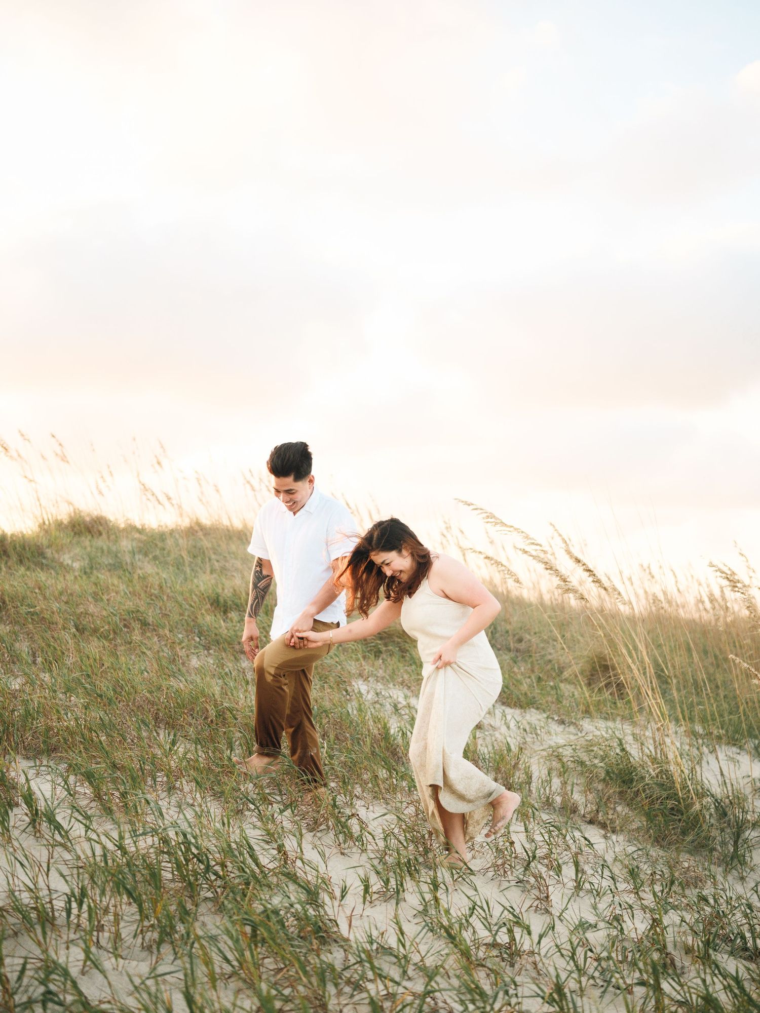 Couple walking through coastal sand dunes with tall beach grass swaying in the breeze at golden hour.