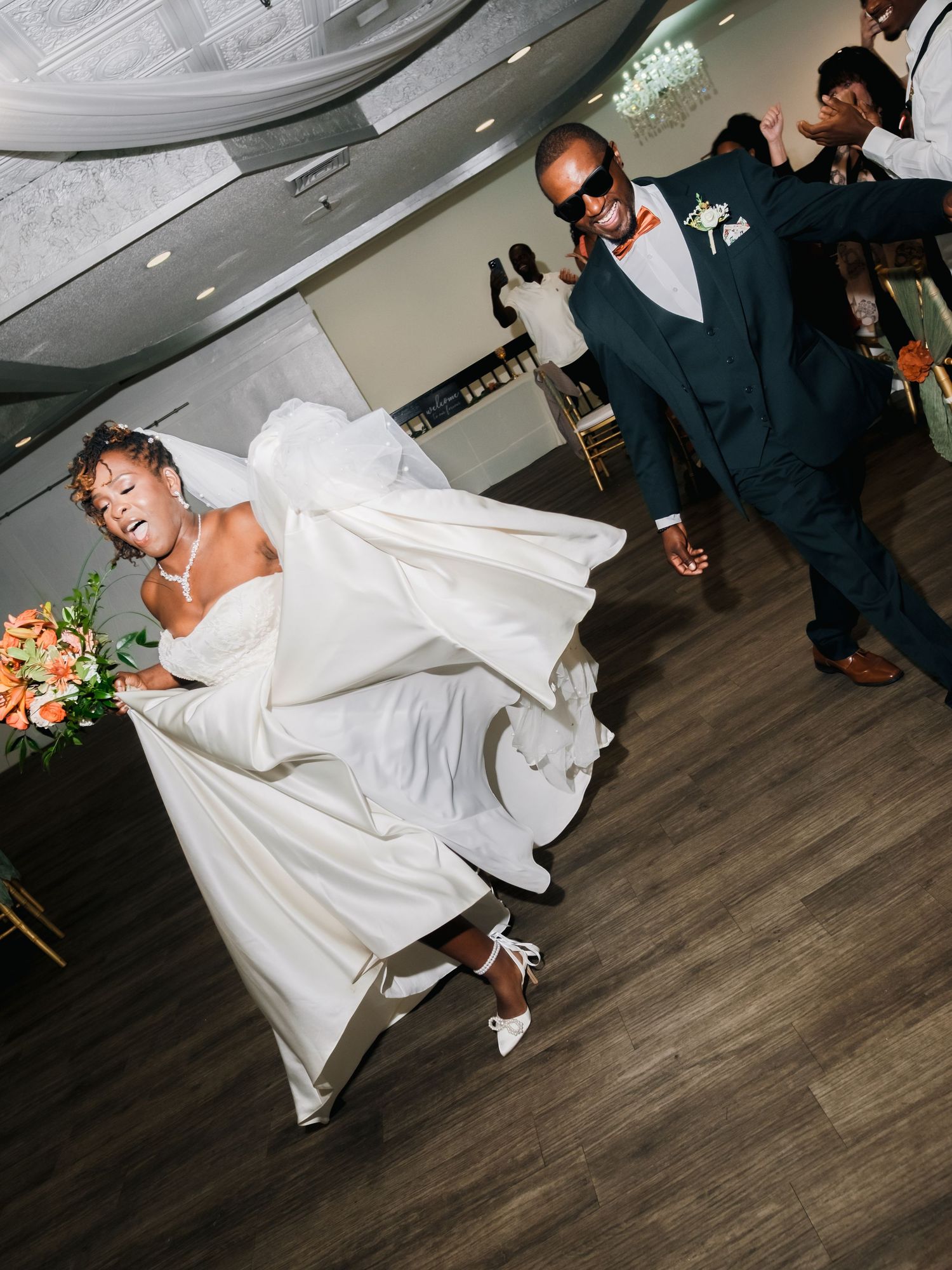 A bride in a flowing white dress performs an energetic dance move on a wooden floor during a wedding reception.