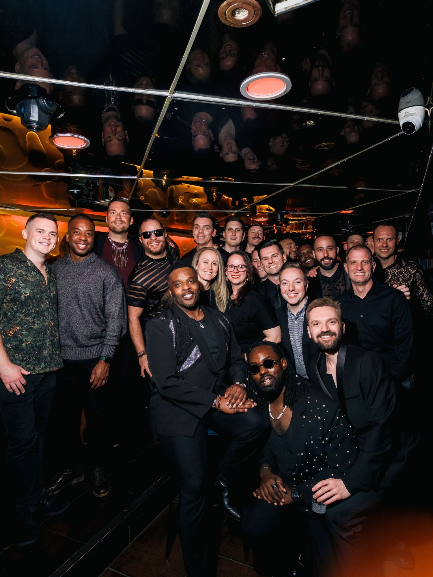 A festive group photo of people gathered in a dimly lit bar or venue with overhead lighting.