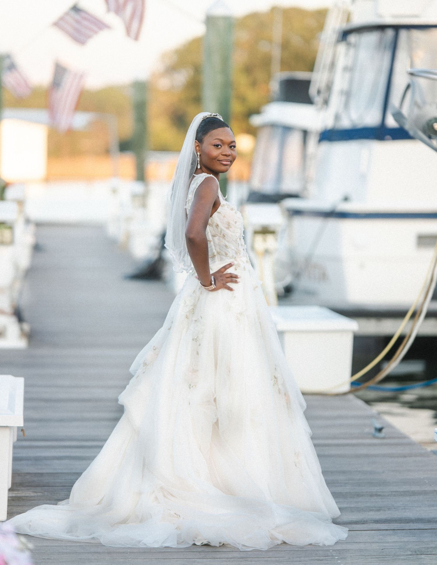A bride in an elegant white lace wedding gown poses on a dock with boats in the marina behind her during golden hour.