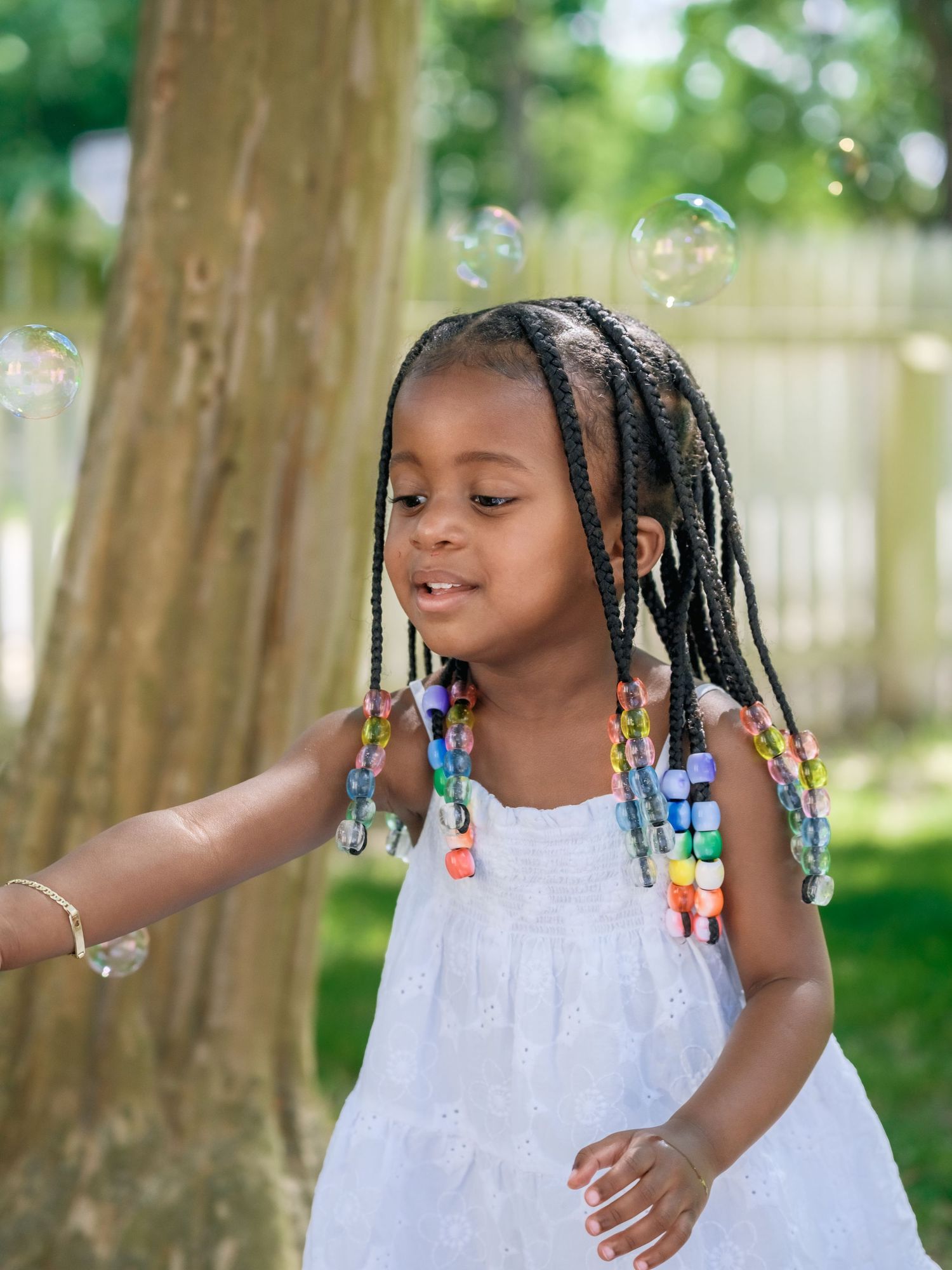 A child in a white dress plays with soap bubbles in a sunny backyard, wearing colorful beaded braids.