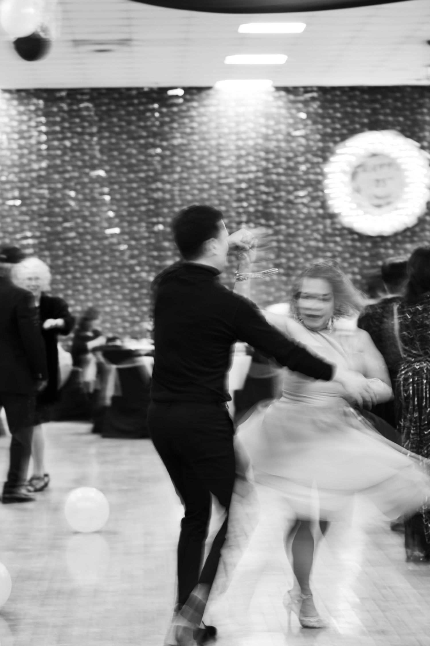Dancers in a ballroom spin and twirl in a blurred black and white photo with a brick wall backdrop.