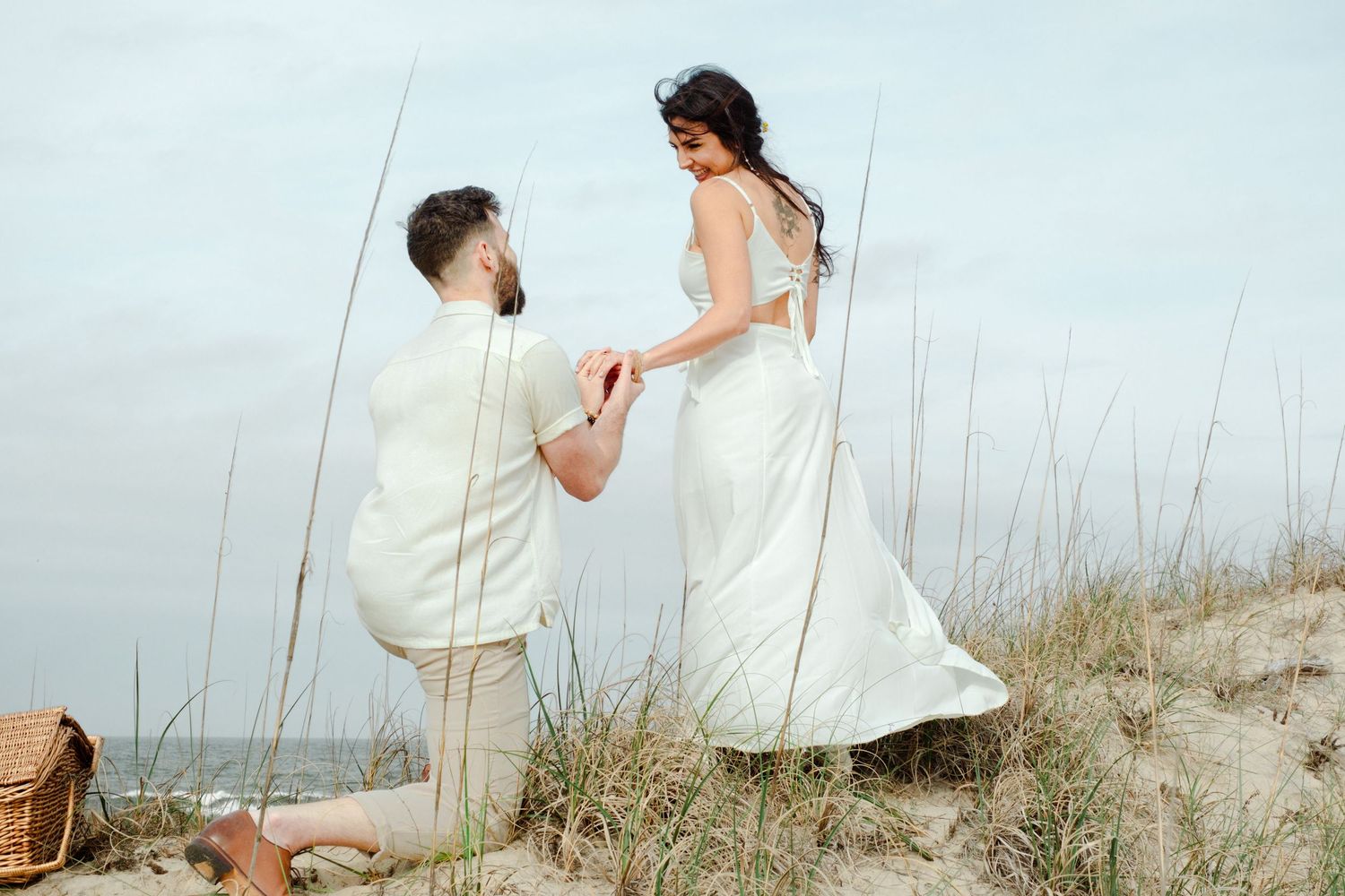Couple in matching white outfits during a romantic beach proposal with a picnic basket on a sandy dune.