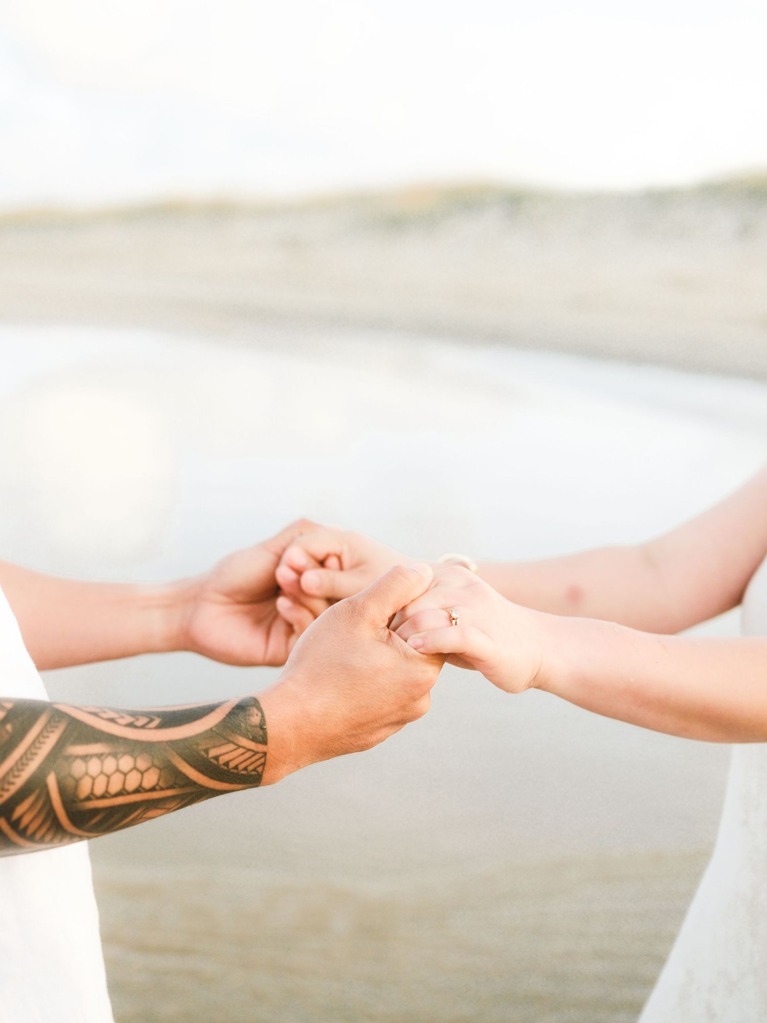 Close up of hands holding each other with one arm showing tribal tattoo design against soft neutral background.