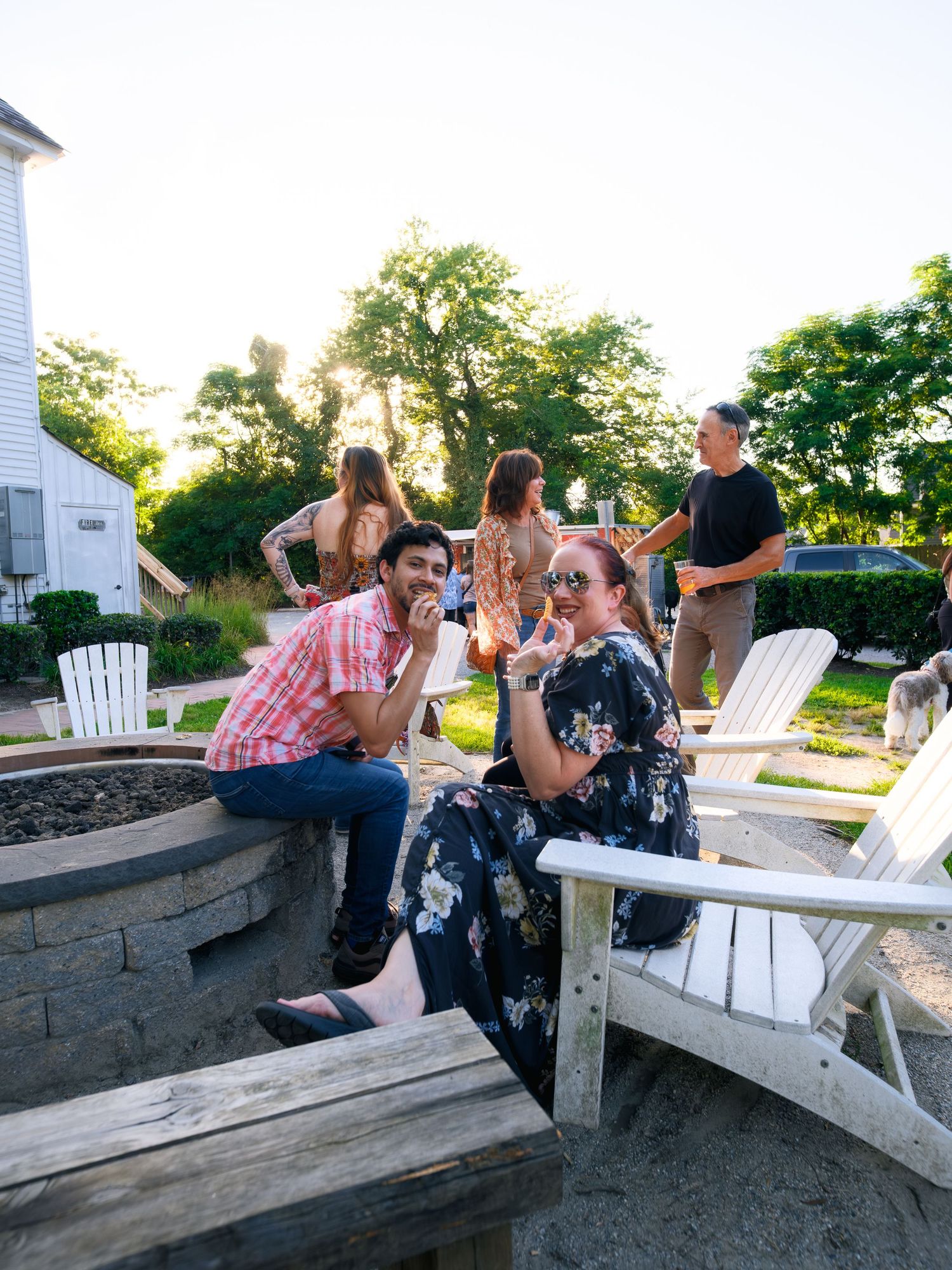 People enjoying a summer evening gathering around a stone fire pit with white Adirondack chairs in an outdoor backyard setting.