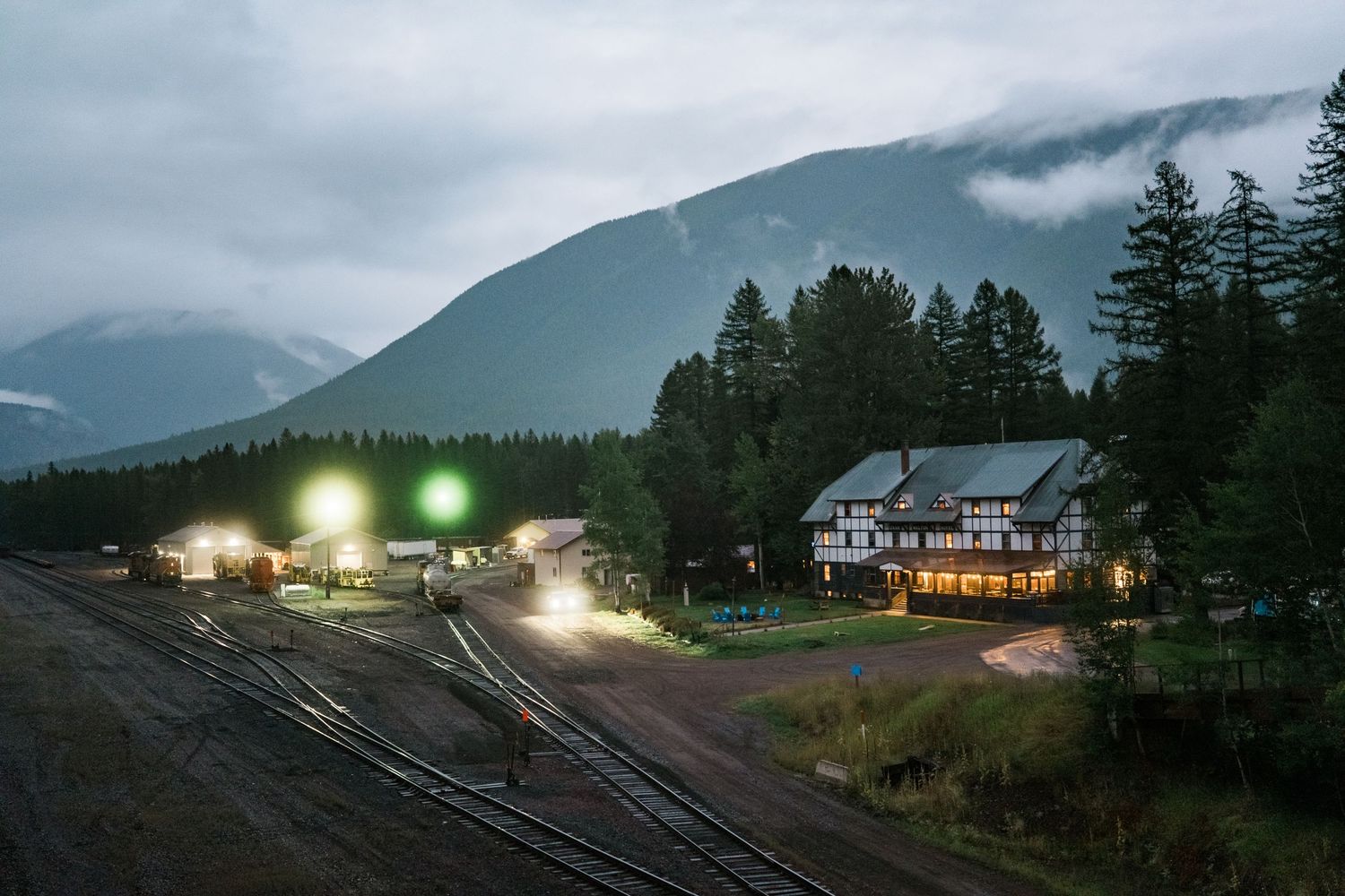 A series of mountain railroad tracks at dusk with glowing lights from buildings and trains in the misty landscape.