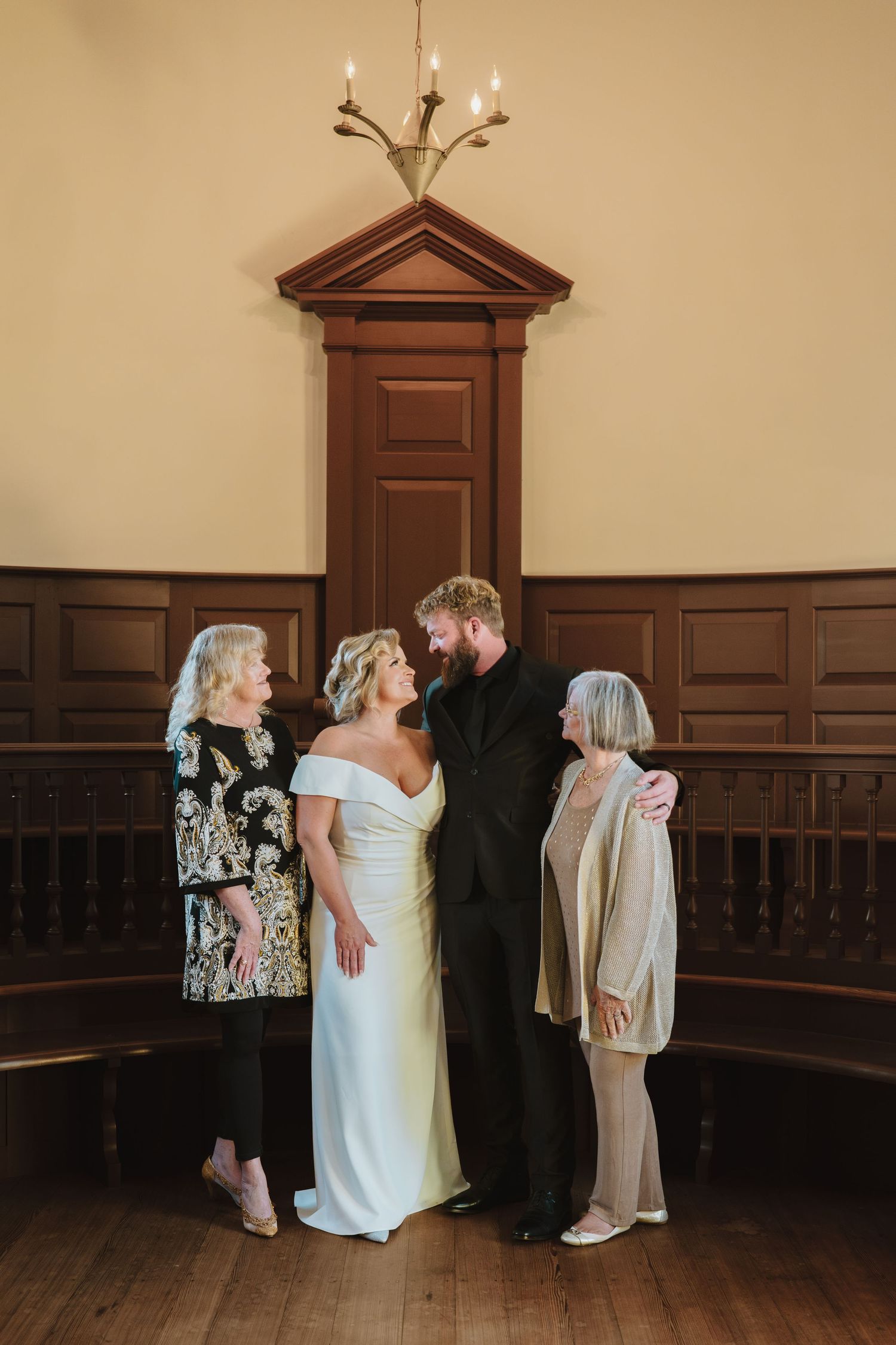 Two people in wedding attire share an intimate moment against a wood-paneled wall in a classic interior space.
