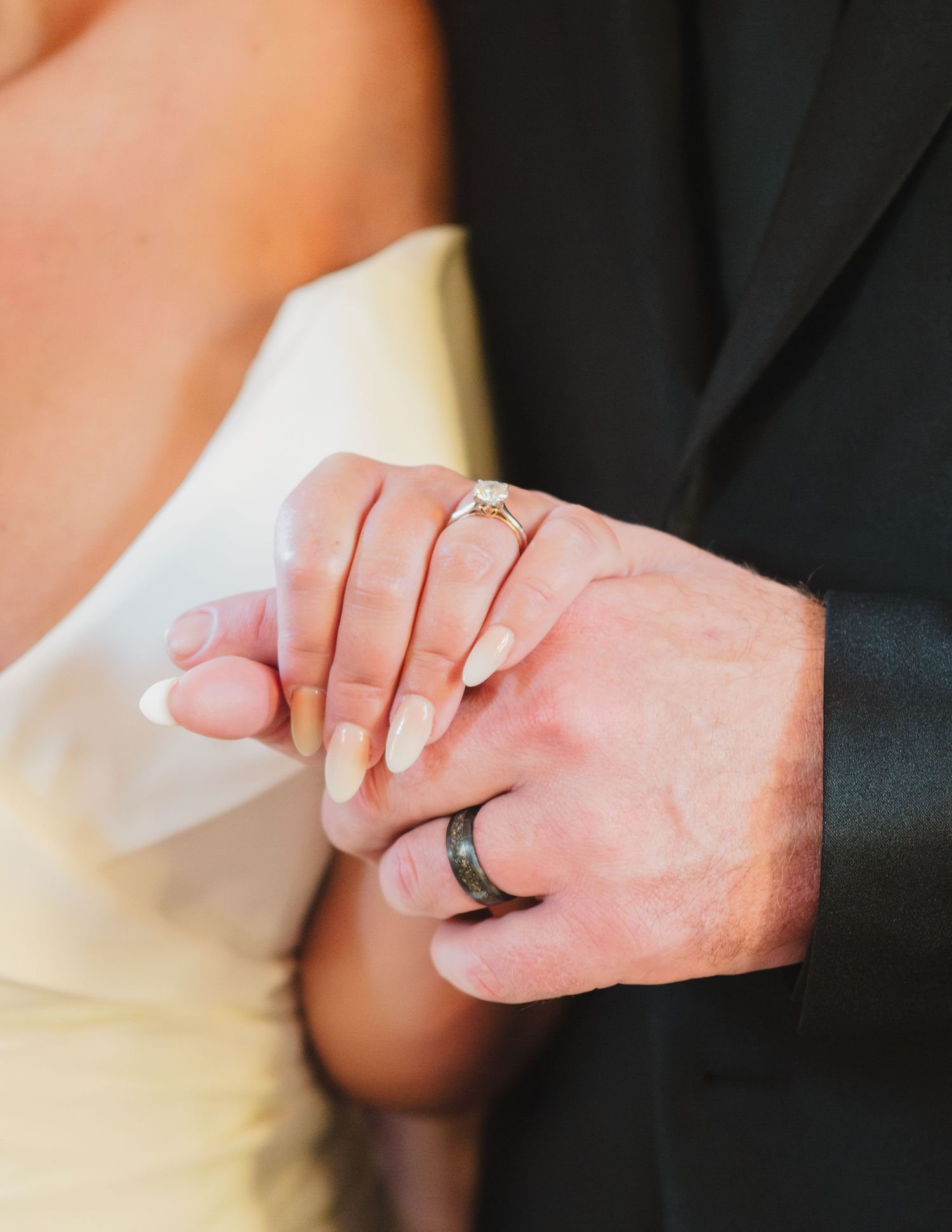 Close-up of newlywed couple's hands showing off their wedding rings against white and black formal attire.