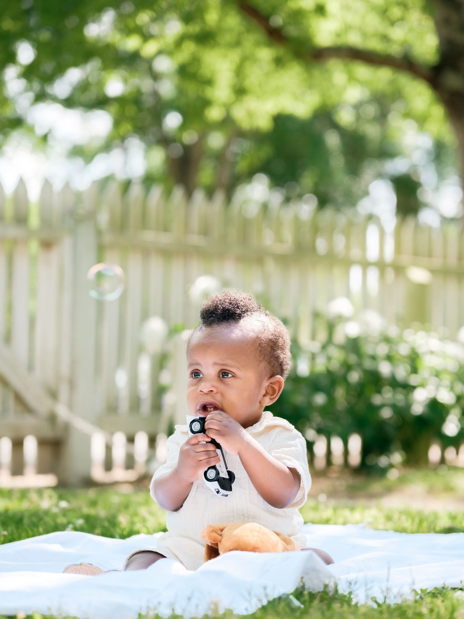 A baby in formal wear sits on a white blanket in a sunny backyard with a white picket fence in the background.
