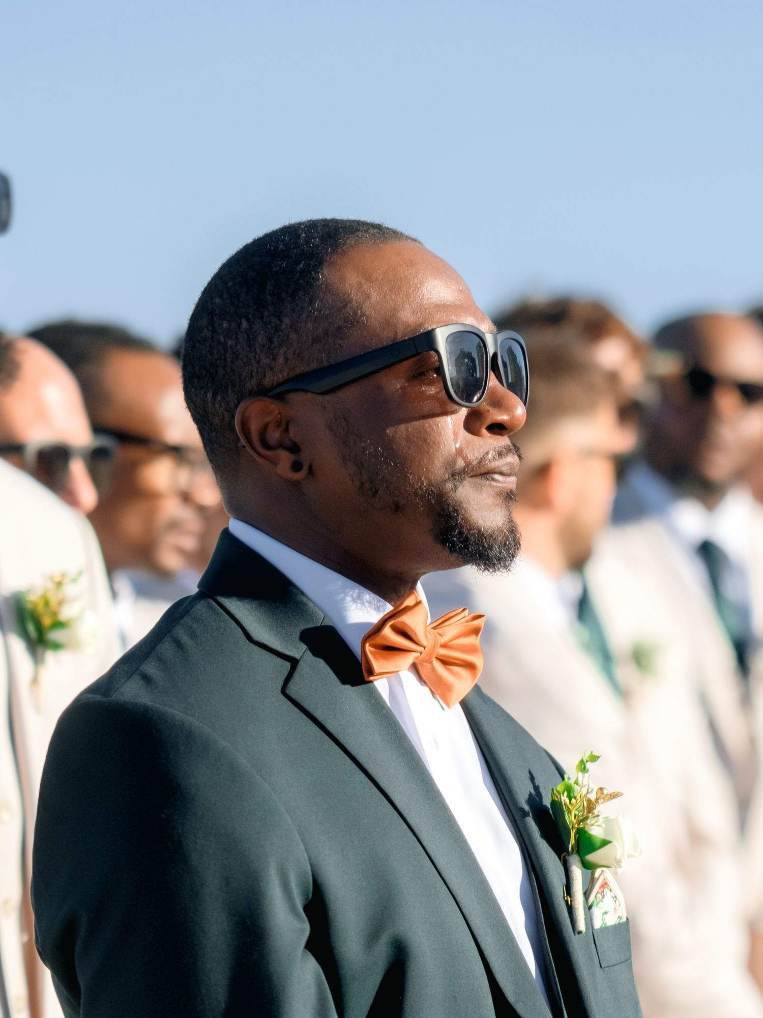 Groomsmen in dark suits and sunglasses stand together during an outdoor beach wedding.