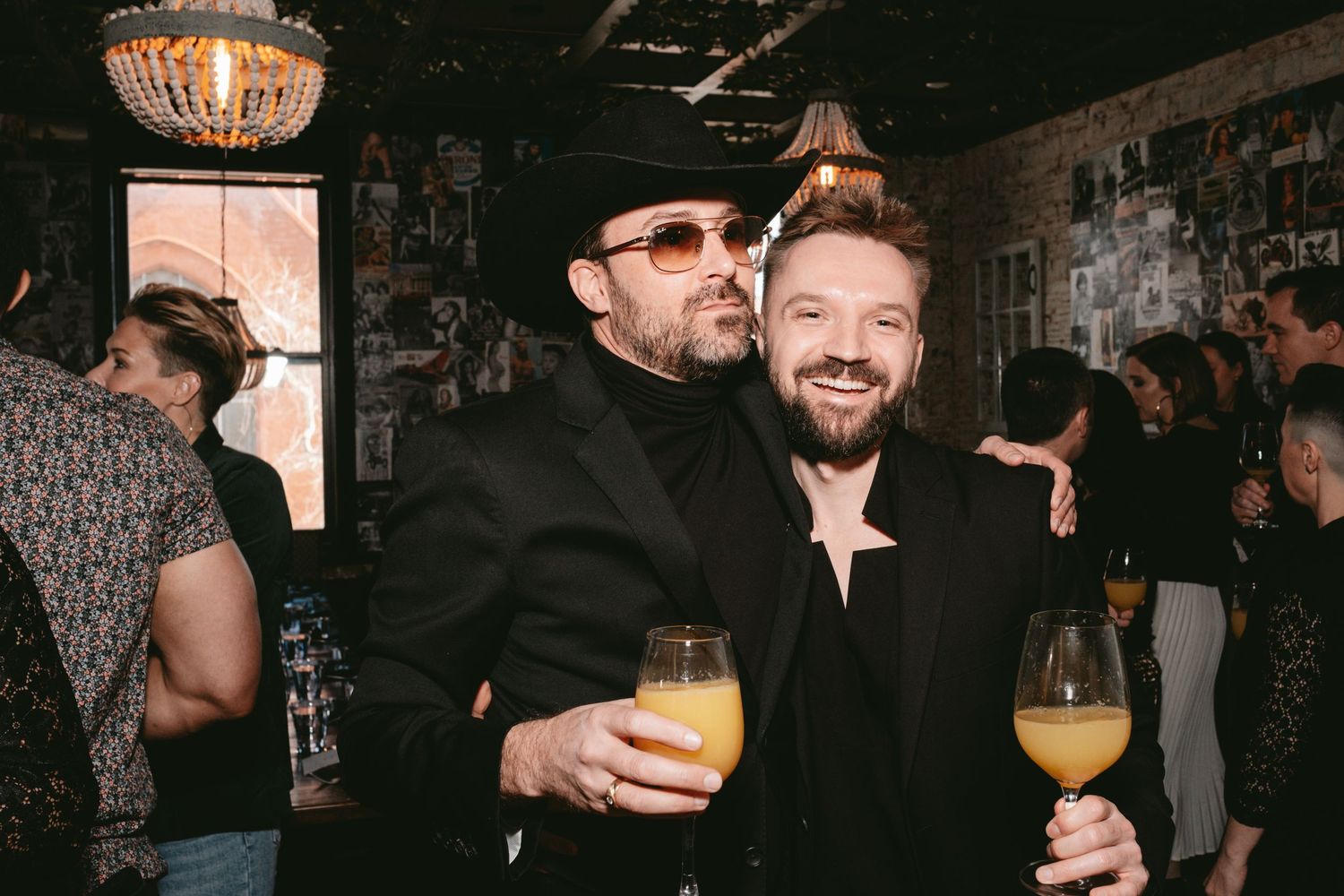 Two smiling friends enjoying drinks together at a dimly lit bar with a chandelier overhead and other patrons in background.