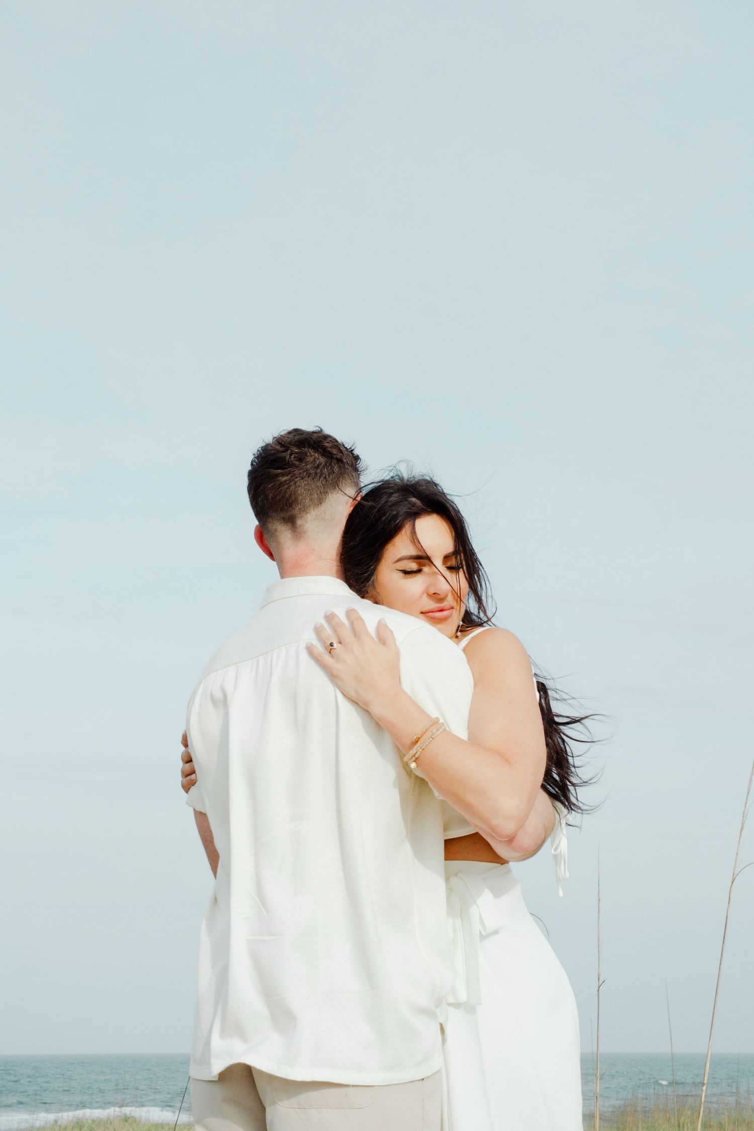 A couple embraces on a beach during a romantic coastal photo session, dressed in white against a soft blue sky.