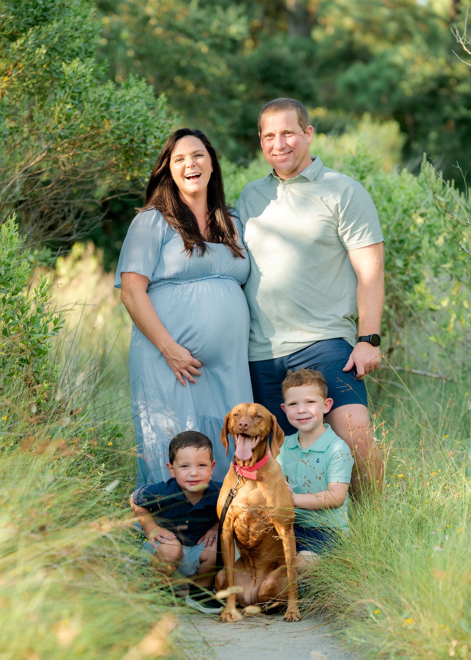 Family portrait with dog in nature setting featuring expecting mother in blue dress.