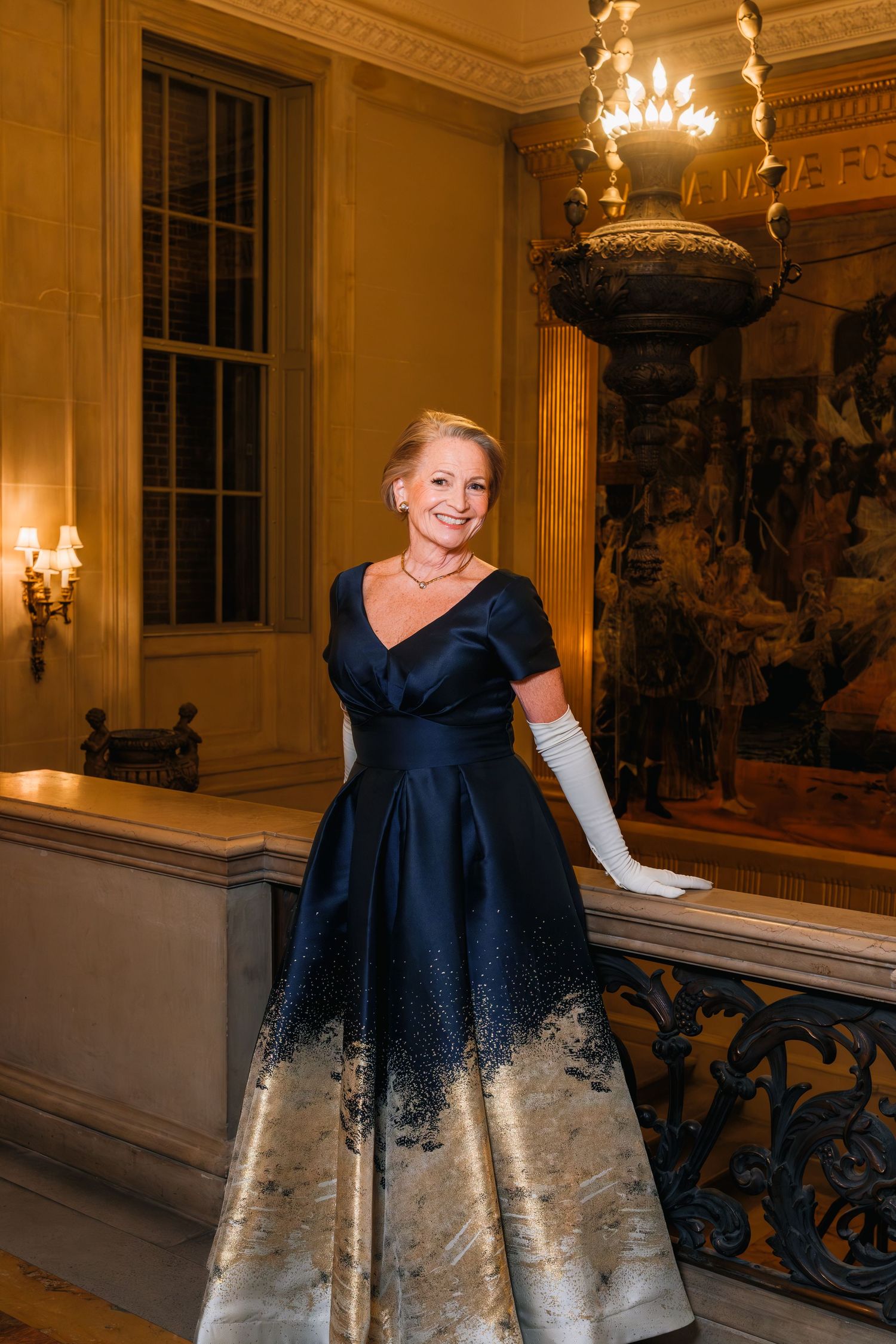 Woman in elegant navy blue and silver ball gown poses in ornate ballroom with chandeliers and decorative railings.