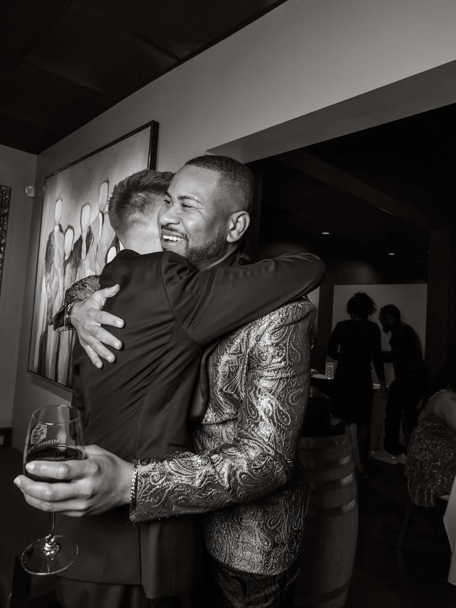 Two people share a warm embrace at an event while holding wine glasses.