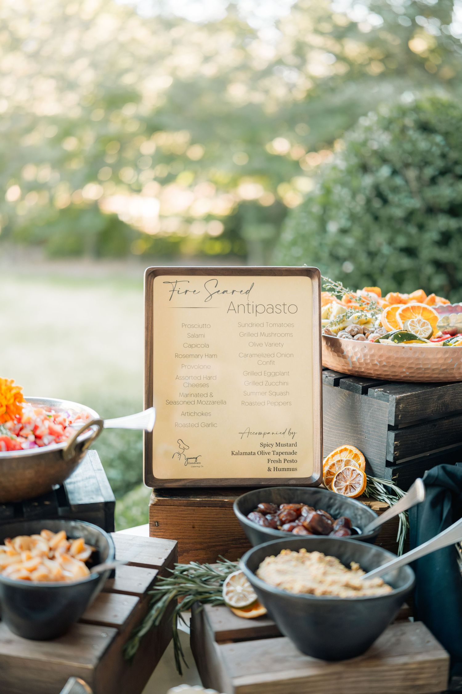 Appetizer station at an outdoor catering event with rustic wooden menus and black serving bowls displaying antipasto dishes.