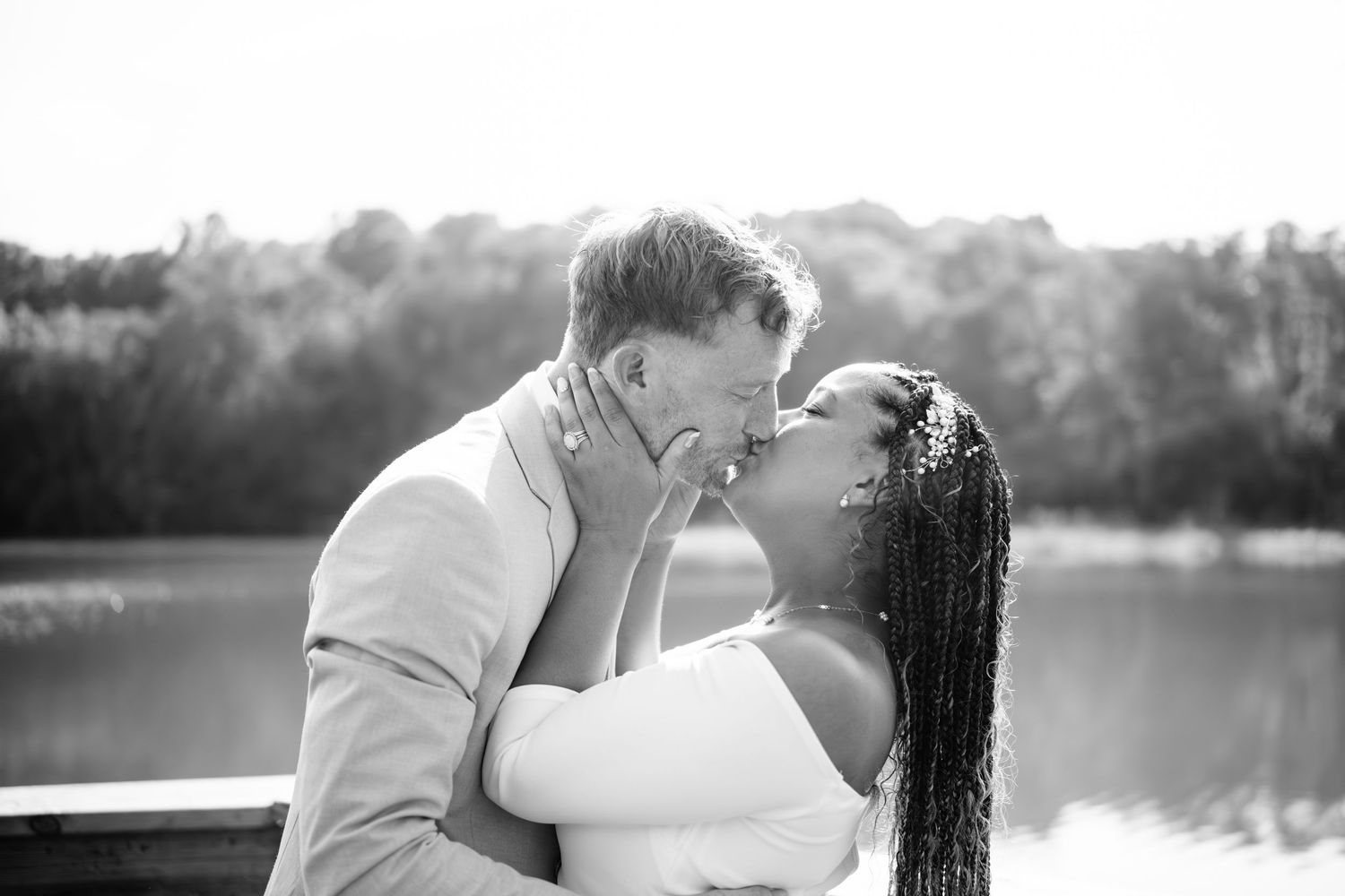 Black and white romantic couple embracing and kissing by a serene lake with trees in the background.