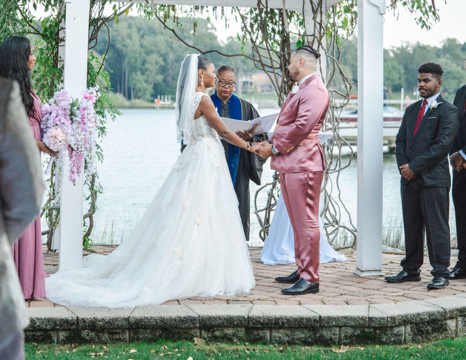 An outdoor wedding ceremony takes place under floral decorations beside a tranquil waterfront setting on a sunny day.