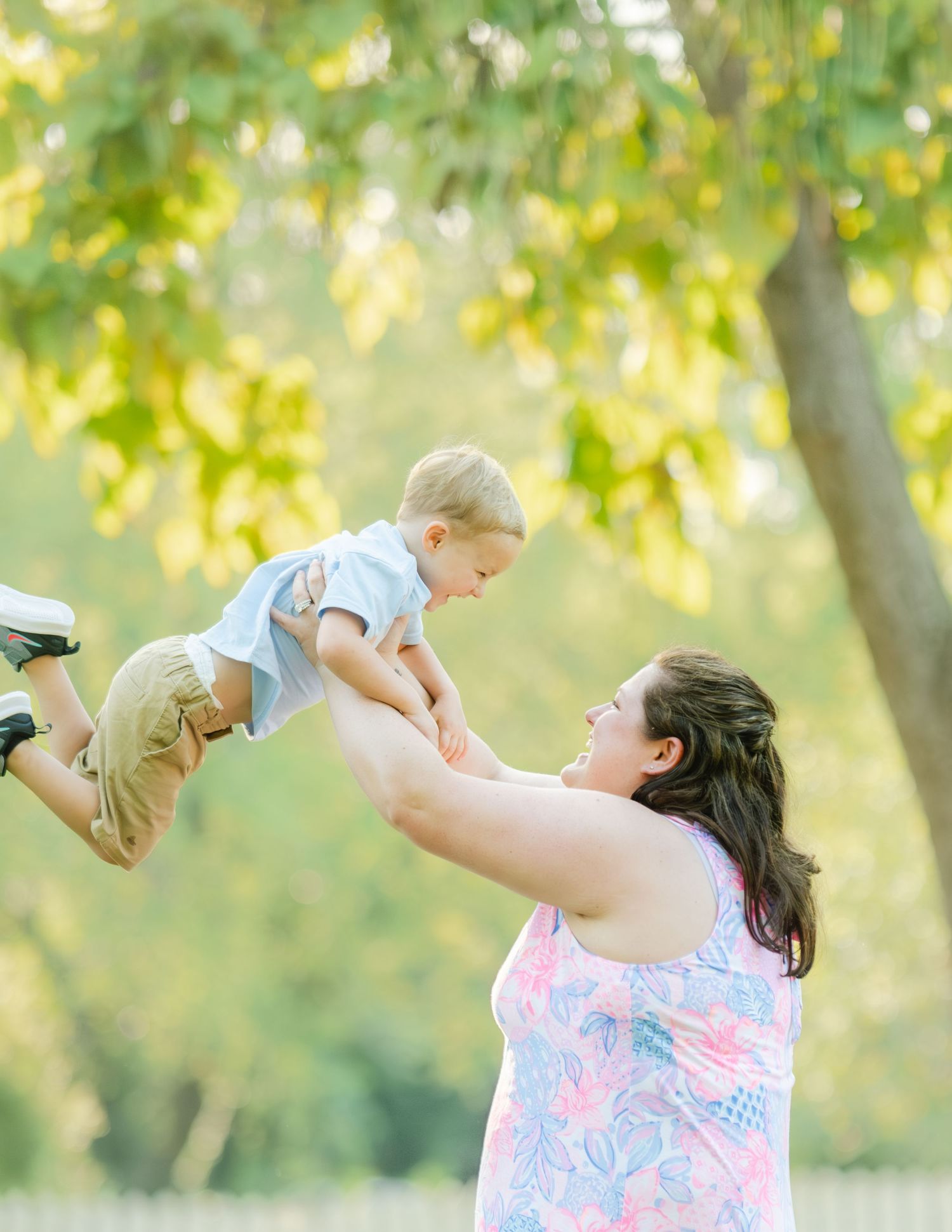 A woman in a floral dress lifts and plays with a young child in a sunny outdoor setting with green trees in the background.