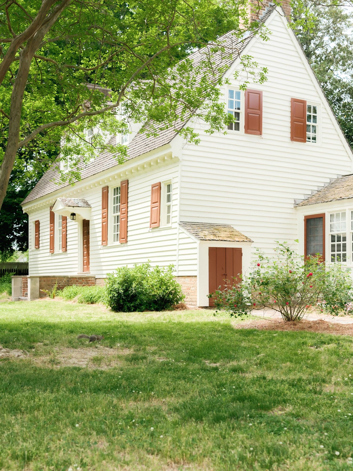 Five connected images show different angles of a traditional white Cape Cod style house with red shutters on a grassy slope.