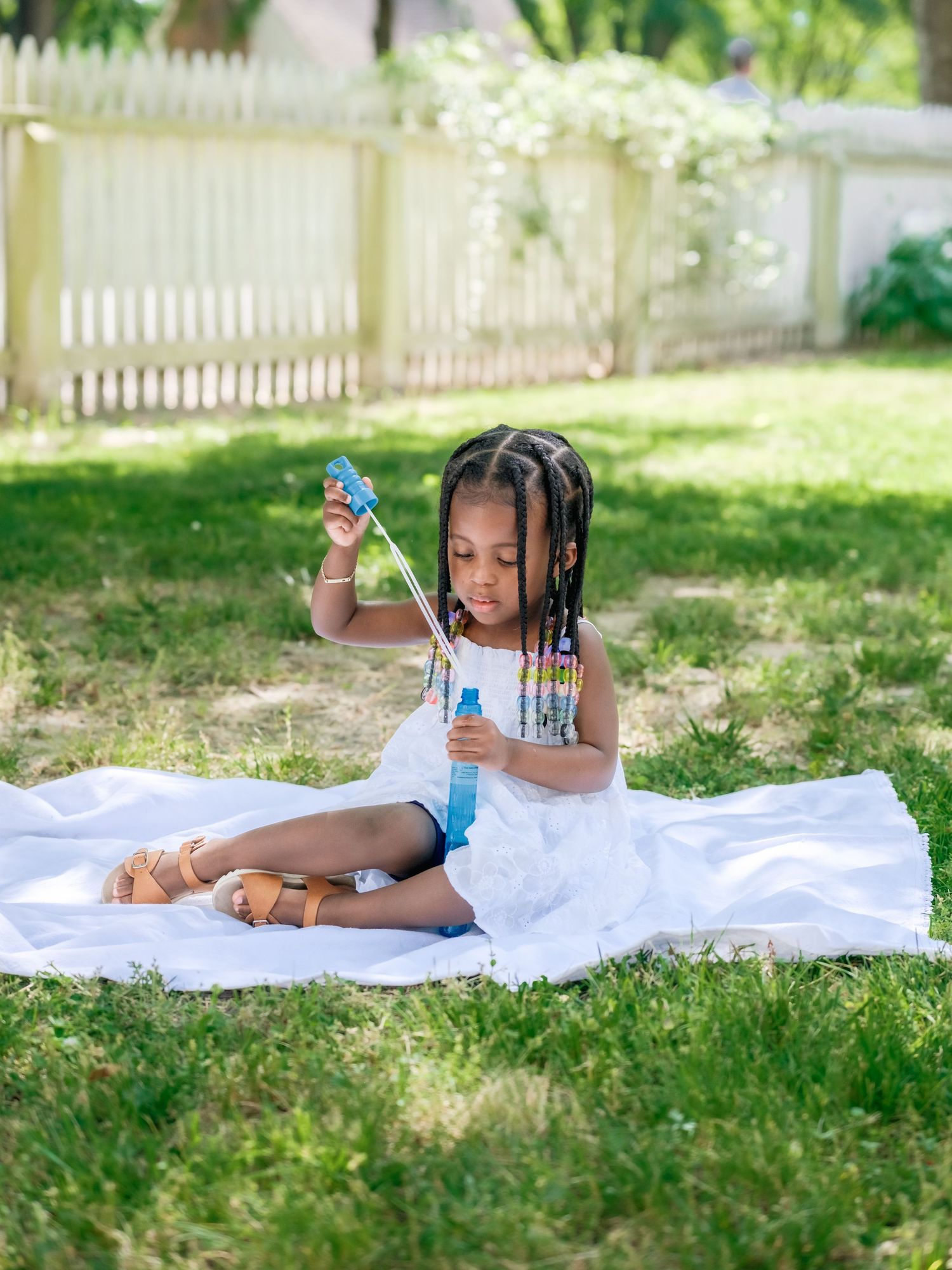 A young child enjoys reading a storybook while laying on a white blanket in a sunny backyard garden.