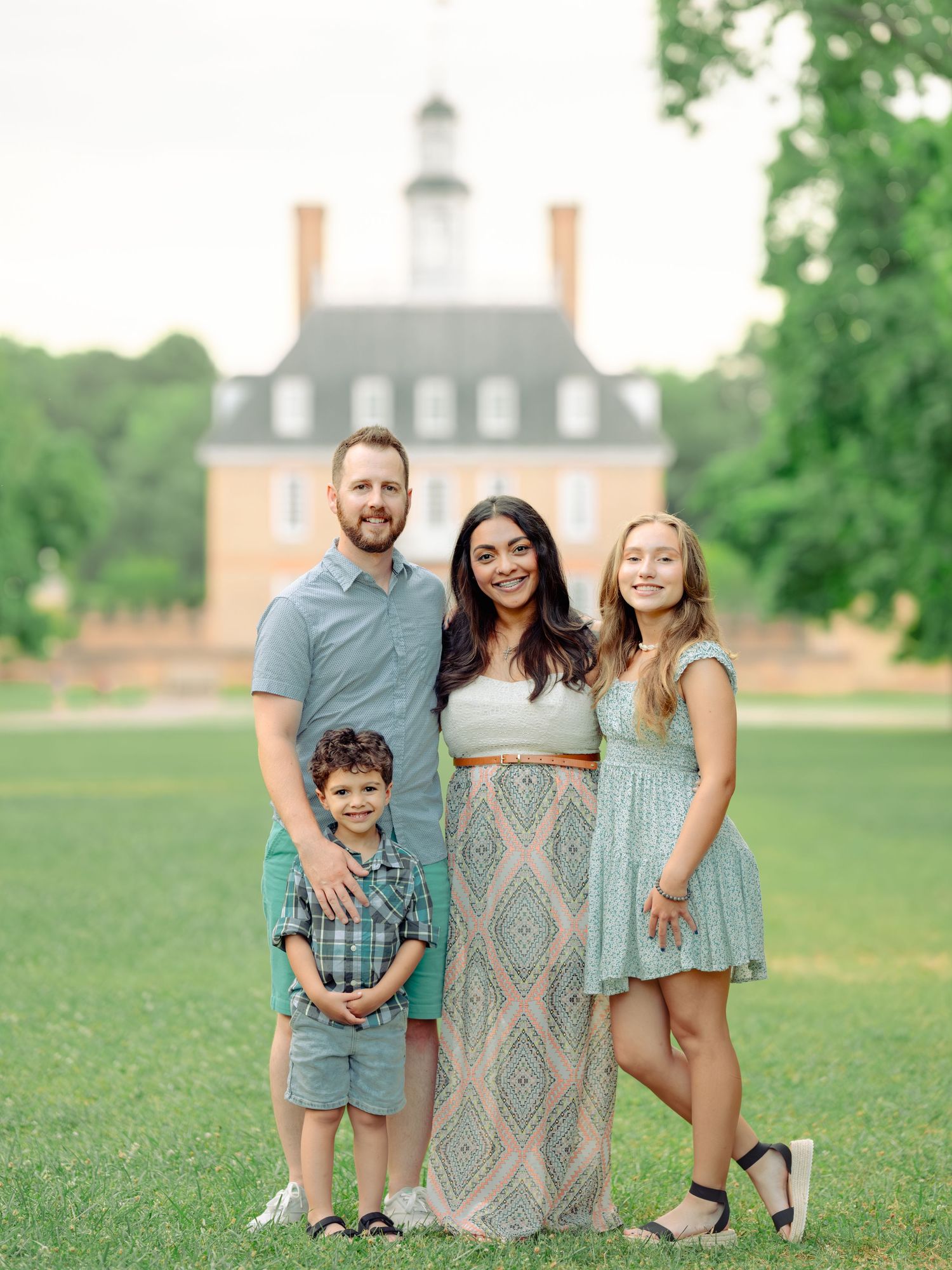 A family wearing coordinating blue and gray outfits poses together on a grassy lawn with brick buildings in the background.